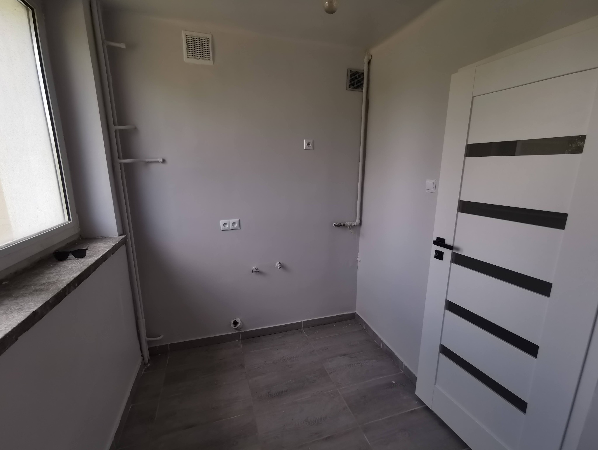 Szare wnętrze w trakcie prac wykończeniowych: widoczne rury, puszki elektryczne, gładkie ściany, szara podłoga imitująca drewno oraz białe drzwi z poziomymi, czarnymi paskami. Na parapecie leżą...