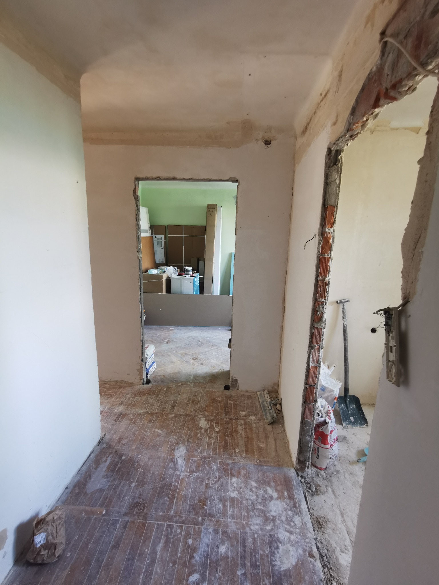 Remont mieszkania w Warszawie: Widok na korytarz z usuniętymi futrynami i odkrytą cegłą, przygotowany do dalszych prac wykończeniowych, z widoczną szpachlą i workiem gipsu.