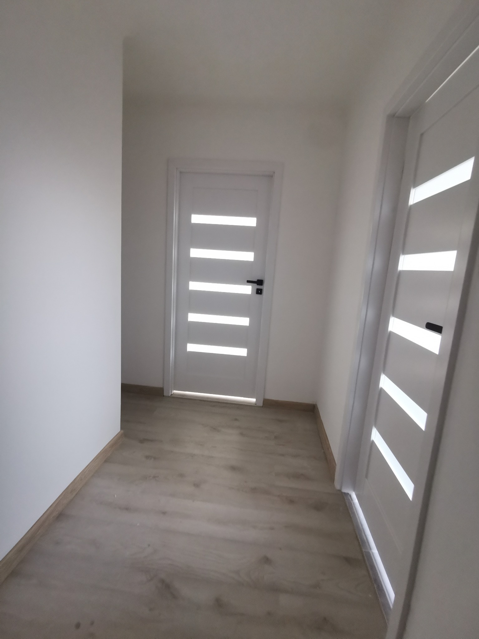 Wykończony korytarz z dwiema białymi, nowoczesnymi drzwiami z poziomymi przeszkleniami i panelami podłogowymi w odcieniach beżu.