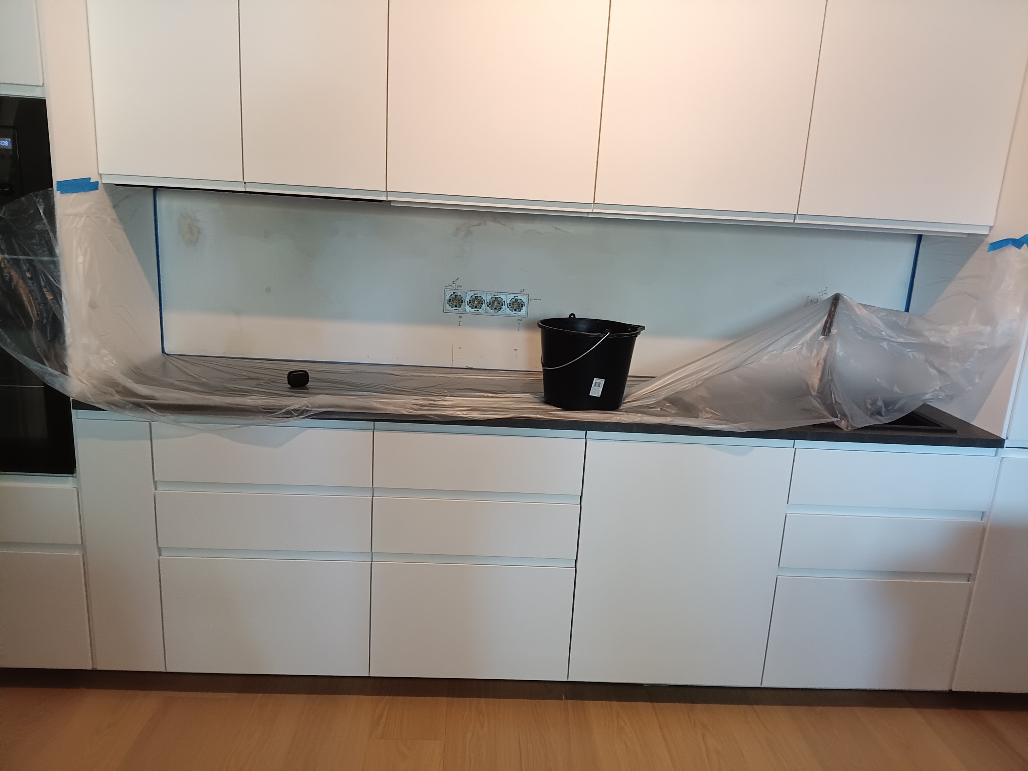 Przygotowanie do malowania ściany w kuchni: zabezpieczony blat folią, widoczne gniazdka elektryczne i czarne wiadro na blacie.