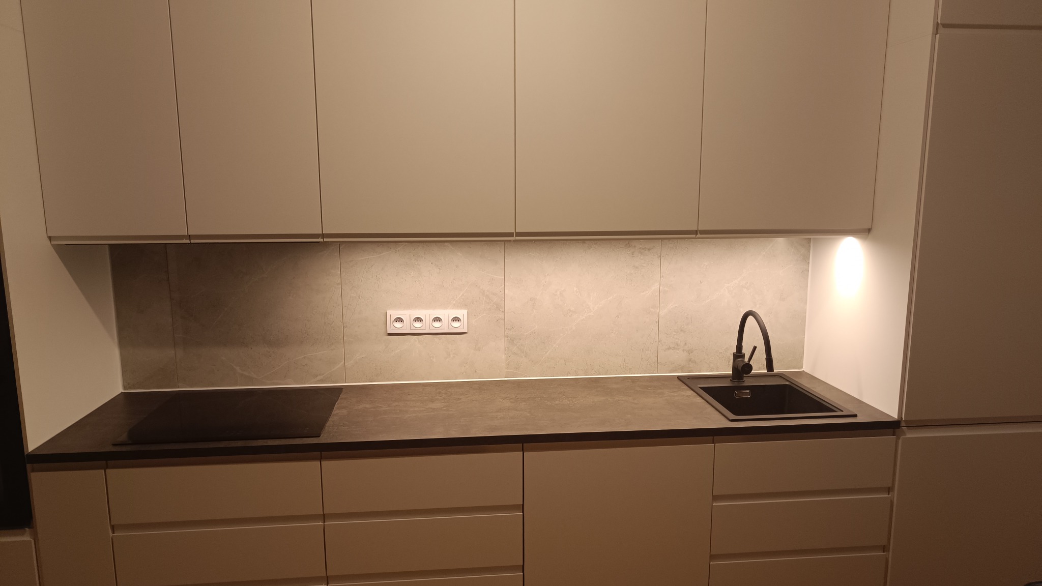 Nowoczesna kuchnia z szarymi frontami szafek, ciemnym blatem i czarną armaturą, oświetlona listwą LED pod szafkami wiszącymi, gniazdka elektryczne na tle płytek imitujących marmur.