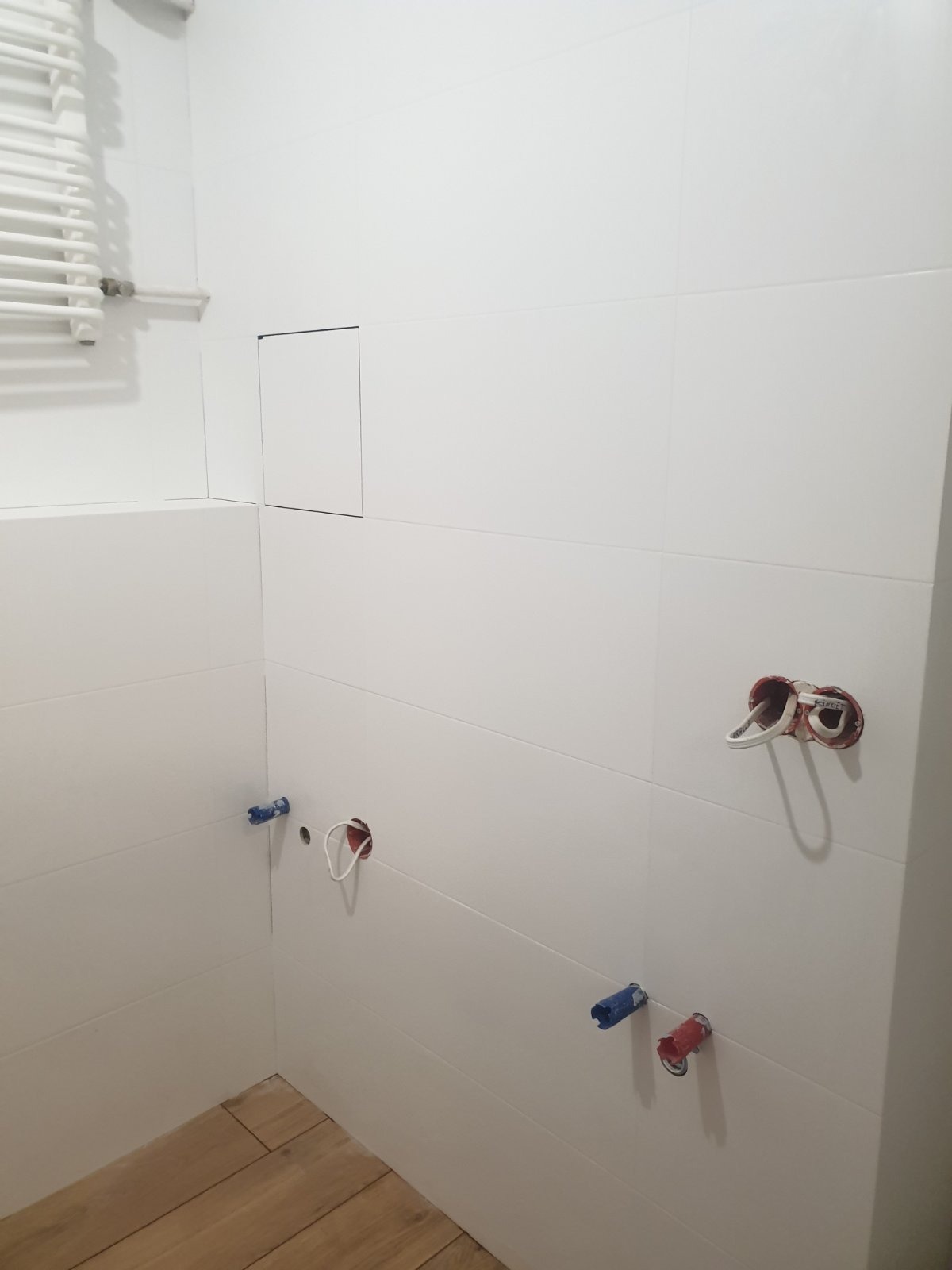 Białe płytki ułożone na ścianach w łazience w Warszawie, widoczne otwory na instalacje wodne i elektryczne, przygotowane do dalszych prac wykończeniowych.