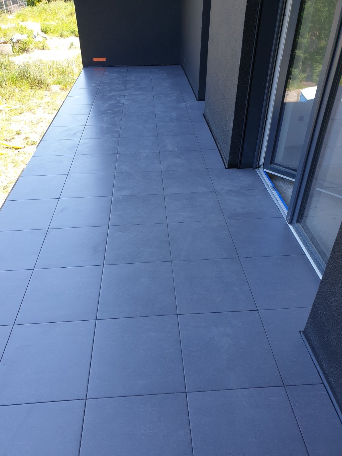 Szare, matowe płytki gresowe ułożone na balkonie z widokiem na zieleń, z ciemnoszarą elewacją i częściowo otwartymi drzwiami balkonowymi.