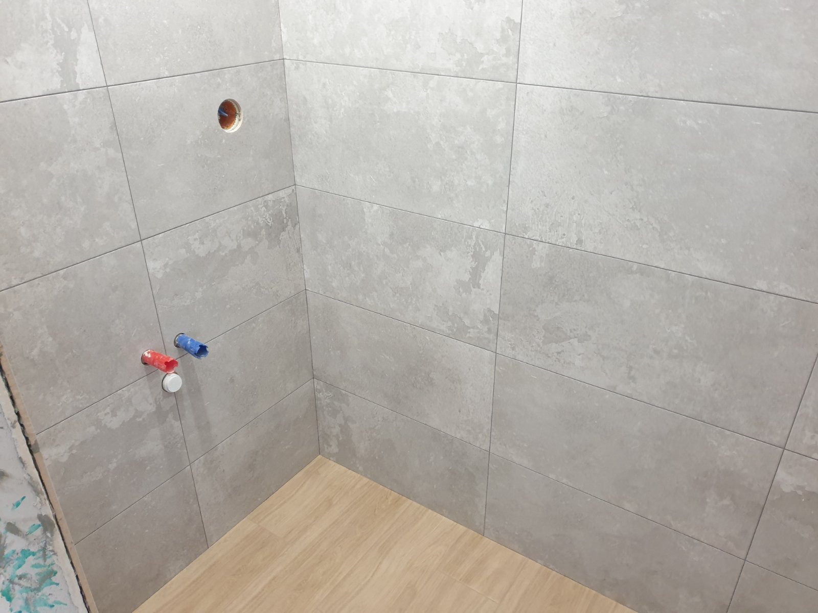 Szare, matowe płytki ceramiczne imitujące kamień na ścianach i podłodze w rogu łazienki, widoczne przyłącza wodne z wystającymi rurkami w kolorze czerwonym i niebieskim oraz otwór na baterię.