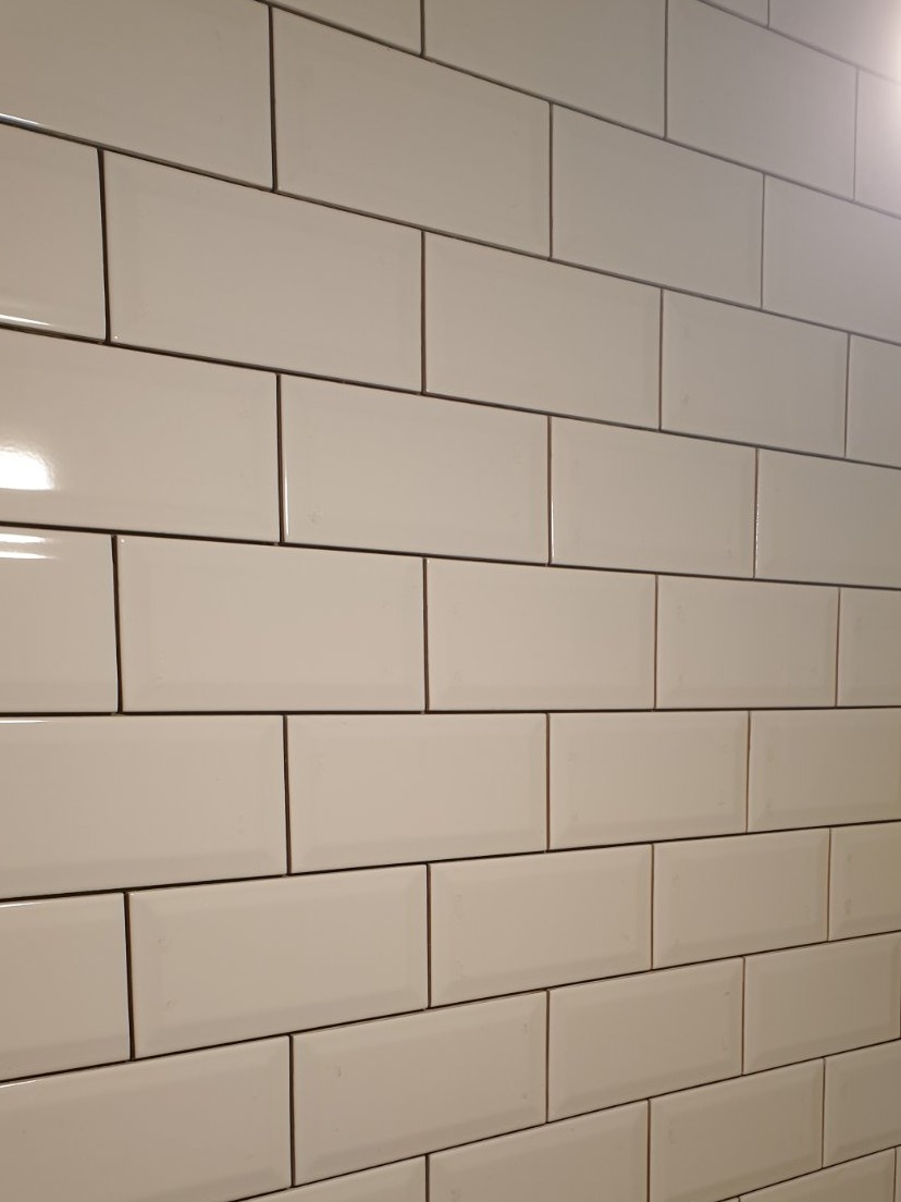 Ściana wyłożona błyszczącymi, kremowymi płytkami ceramicznymi w układzie cegiełki, z ciemnoszarą fugą, ujęcie pod kątem.