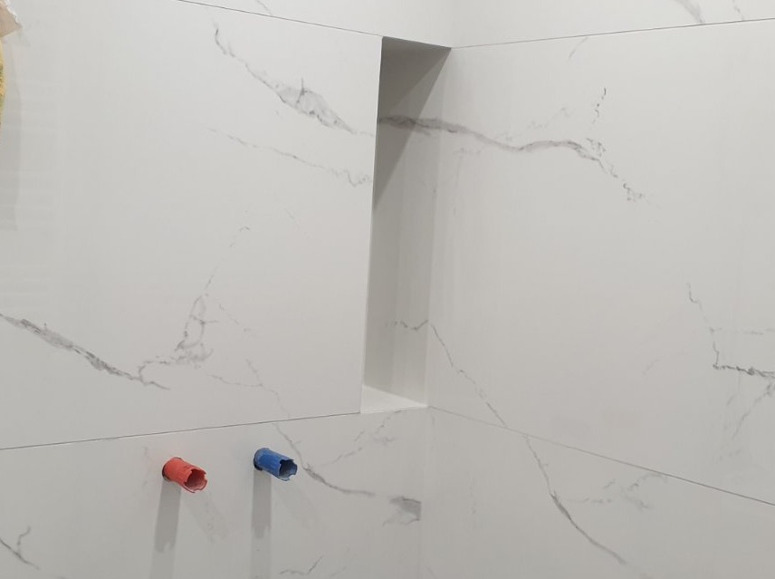 Ściana łazienki wyłożona białymi płytkami imitującymi marmur, z wnęką i wystającymi rurami z zaślepkami w kolorze czerwonym i niebieskim.