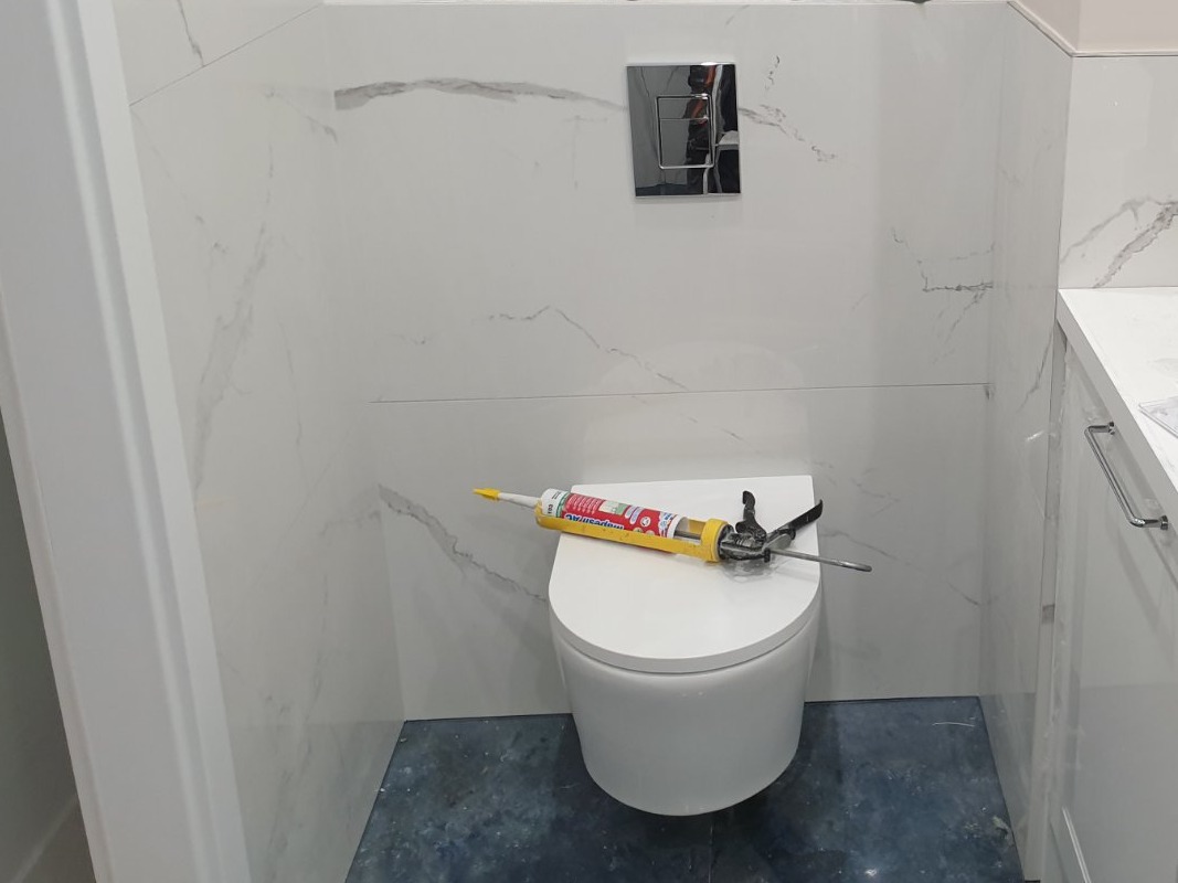 Nowoczesna toaleta wisząca z białymi płytkami imitującymi marmur na ścianach, na desce leży tuba z silikonem i pistolet do silikonu, widoczny chromowany przycisk spłuczki