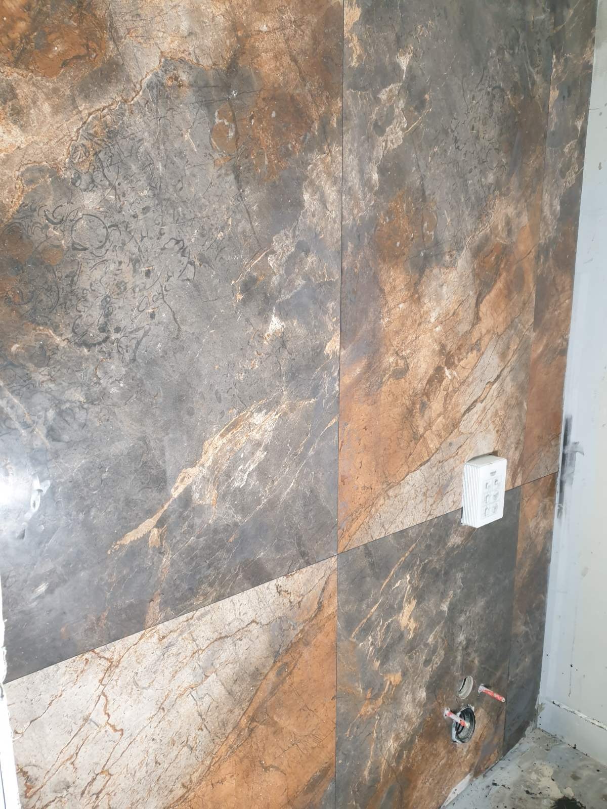 Ściana wyłożona płytkami gresowymi imitującymi marmur w odcieniach szarości, brązu i beżu, z widoczną puszką elektryczną i instalacją hydrauliczną w trakcie montażu.