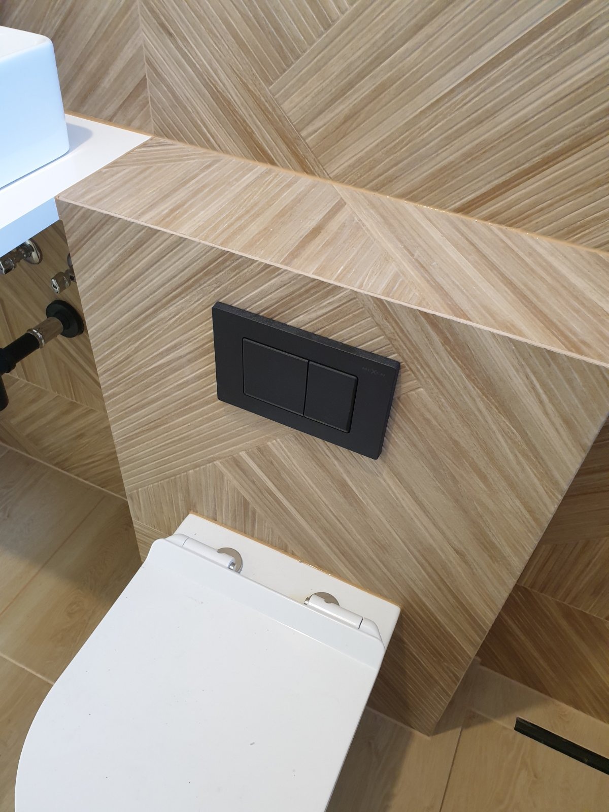 Wisząca toaleta z podtynkowym systemem spłukiwania na tle ściany wyłożonej płytkami w jodełkę, widoczne chromowane zawory.