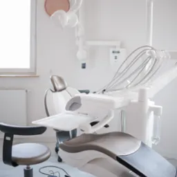 Jasne wnętrze gabinetu z nowoczesnym fotelem dentystycznym w odcieniach bieli i szarości, widoczny zegar na ścianie i marmurowa podłoga.