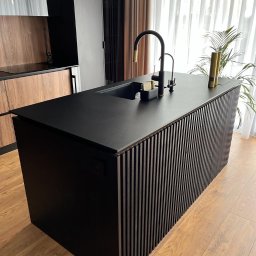 Wood-Mar - Czarna wyspa kuchenna z pionowymi listwami, czarnym blatem i złotą baterią w nowoczesnej kuchni z drewnianymi szafkami w tle.