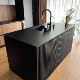 Czarna wyspa kuchenna z pionowymi listwami, czarnym blatem i złotą baterią w nowoczesnej kuchni z drewnianymi szafkami w tle.