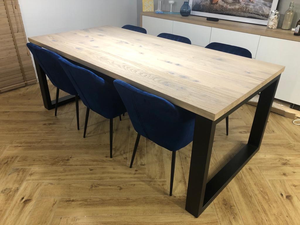 Stół z blatem z jasnego drewna o widocznym usłojeniu, z czarnymi metalowymi nogami i czterema krzesłami obitymi granatowym welurem, ustawiony na podłodze imitującej drewno.