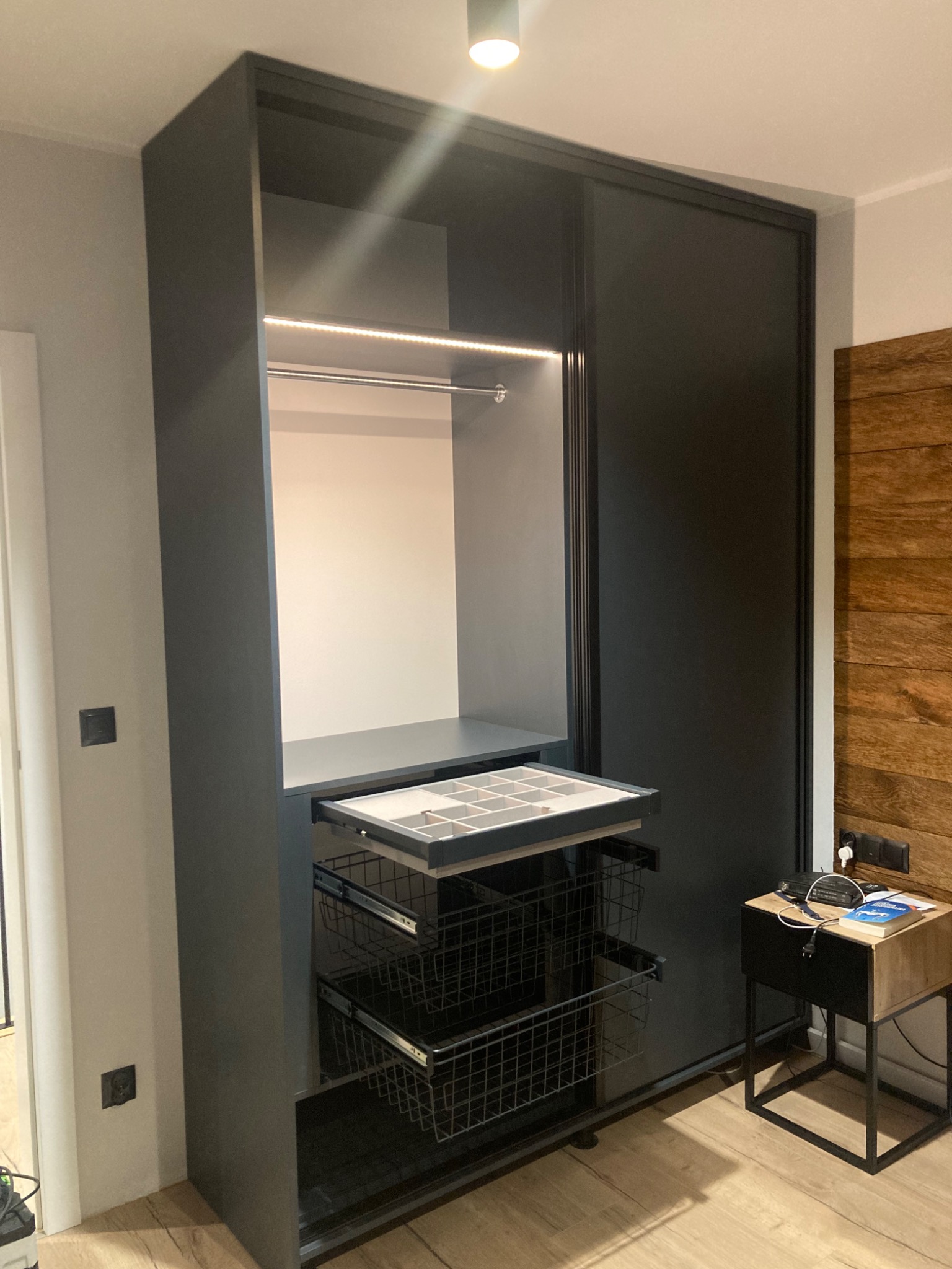 Czarna szafa wnękowa z przesuwnymi drzwiami, oświetleniem LED wewnątrz, wysuwaną półką na drobiazgi i koszami drucianymi, widok z boku na drewnianą ścianę i stolik nocny.