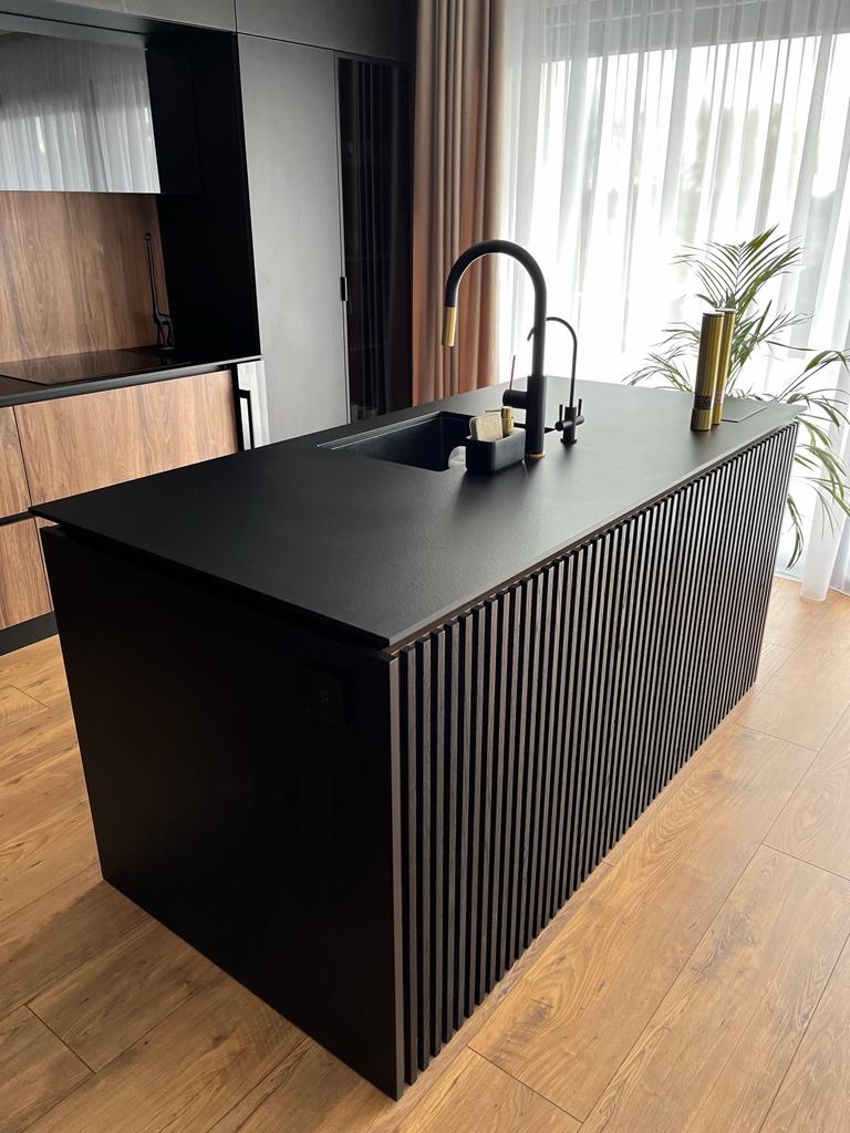 Czarna wyspa kuchenna z pionowymi listwami, czarnym blatem i złotą baterią w nowoczesnej kuchni z drewnianymi szafkami w tle.