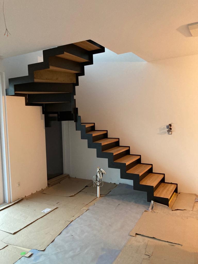 Nowoczesne schody wspornikowe z drewnianymi stopniami i czarną metalową konstrukcją, widoczne w trakcie montażu, z zabezpieczoną podłogą kartonem.