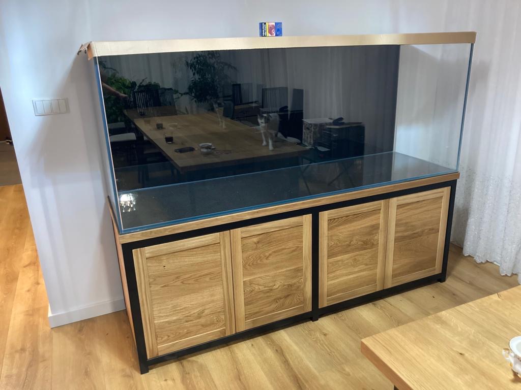 Duże akwarium z drewnianą szafką w stylu rustykalnym, widoczne odbicie wnętrza pomieszczenia w szkle akwarium.