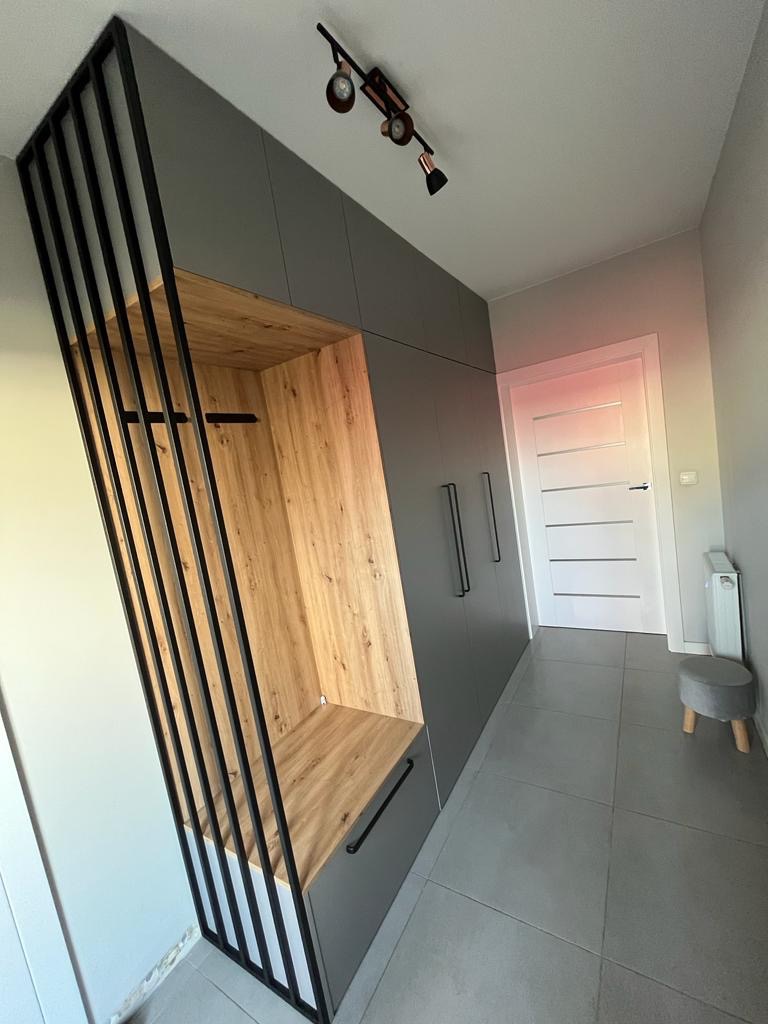 Szara szafa wnękowa z drewnianym wnętrzem i czarną, ażurową konstrukcją, oświetlenie punktowe na suficie, szara podłoga i białe drzwi w tle.