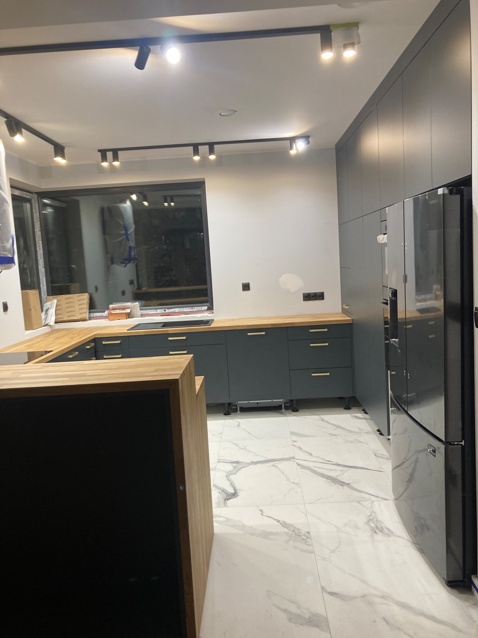 Nowoczesna kuchnia w odcieniach szarości z drewnianym blatem i marmurową podłogą, oświetlona reflektorami sufitowymi.