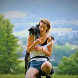 Kobieta kuca na trawie, obejmując czarnego psa na tle rozmytego krajobrazu z drzewami i zabudowaniami.