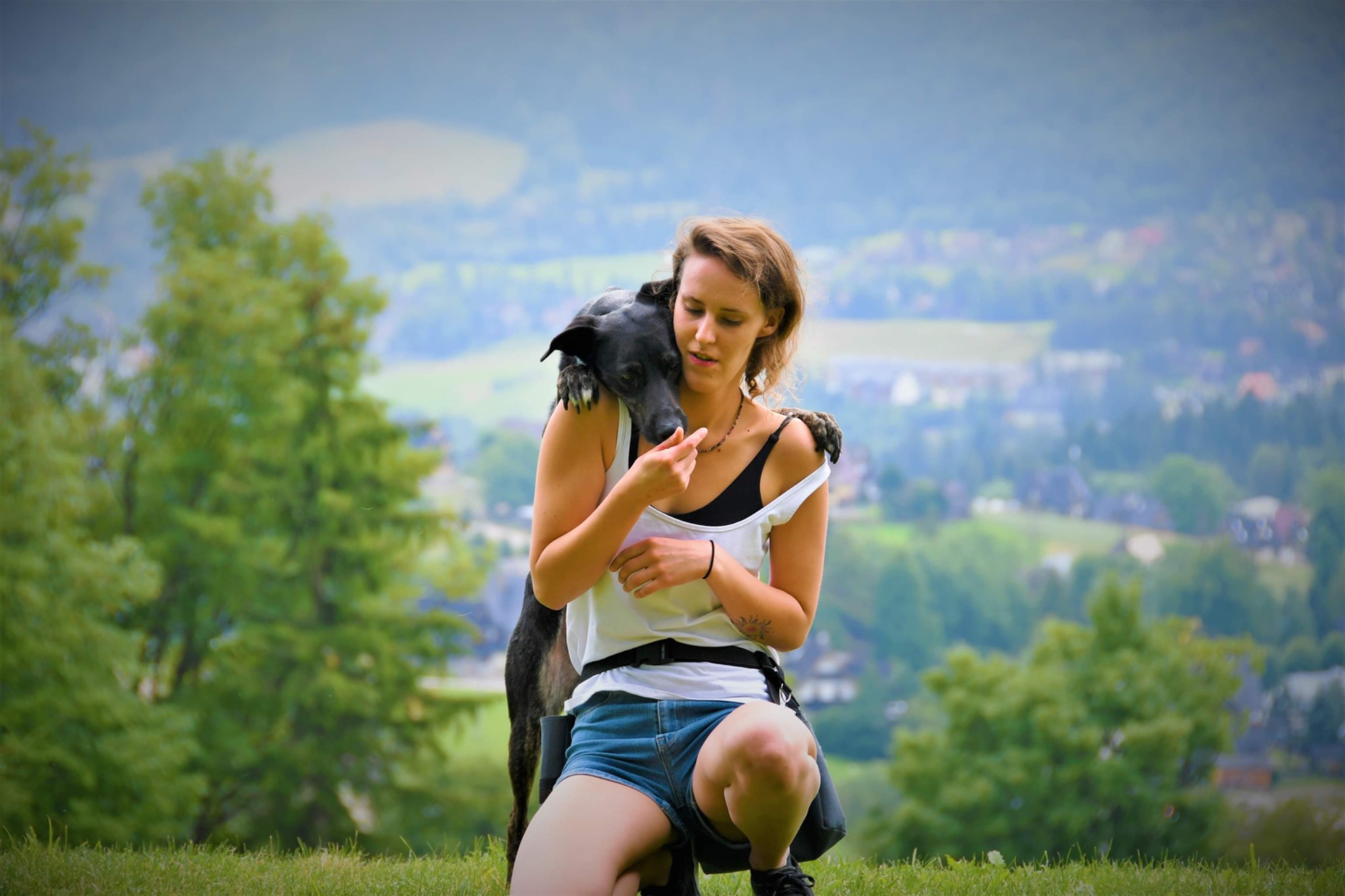 Kobieta kuca na trawie, obejmując czarnego psa na tle rozmytego krajobrazu z drzewami i zabudowaniami.
