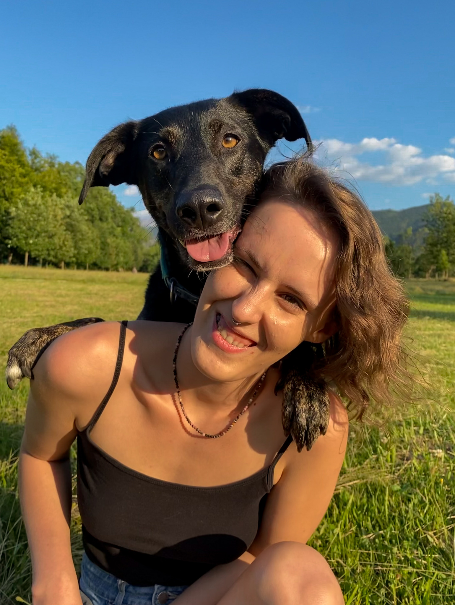Uśmiechnięta kobieta z nosem przekłutym kolczykiem, obejmowana przez czarnego psa na łące w słoneczny dzień.