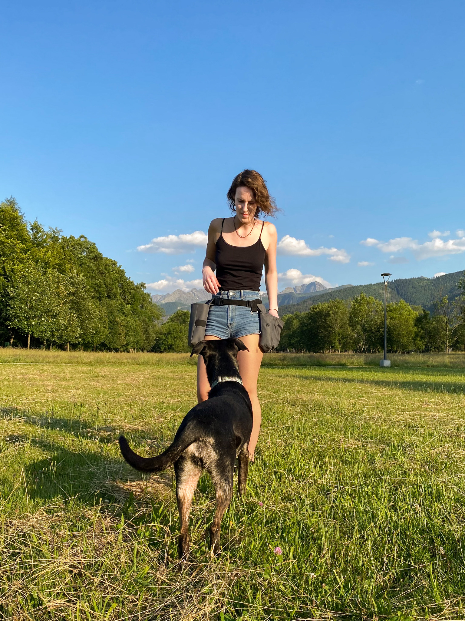 Kobieta w czarnym topie i dżinsowych szortach, z saszetkami na smakołyki, stojąca na trawie z czarnym psem w obroży, góry i drzewa w tle.