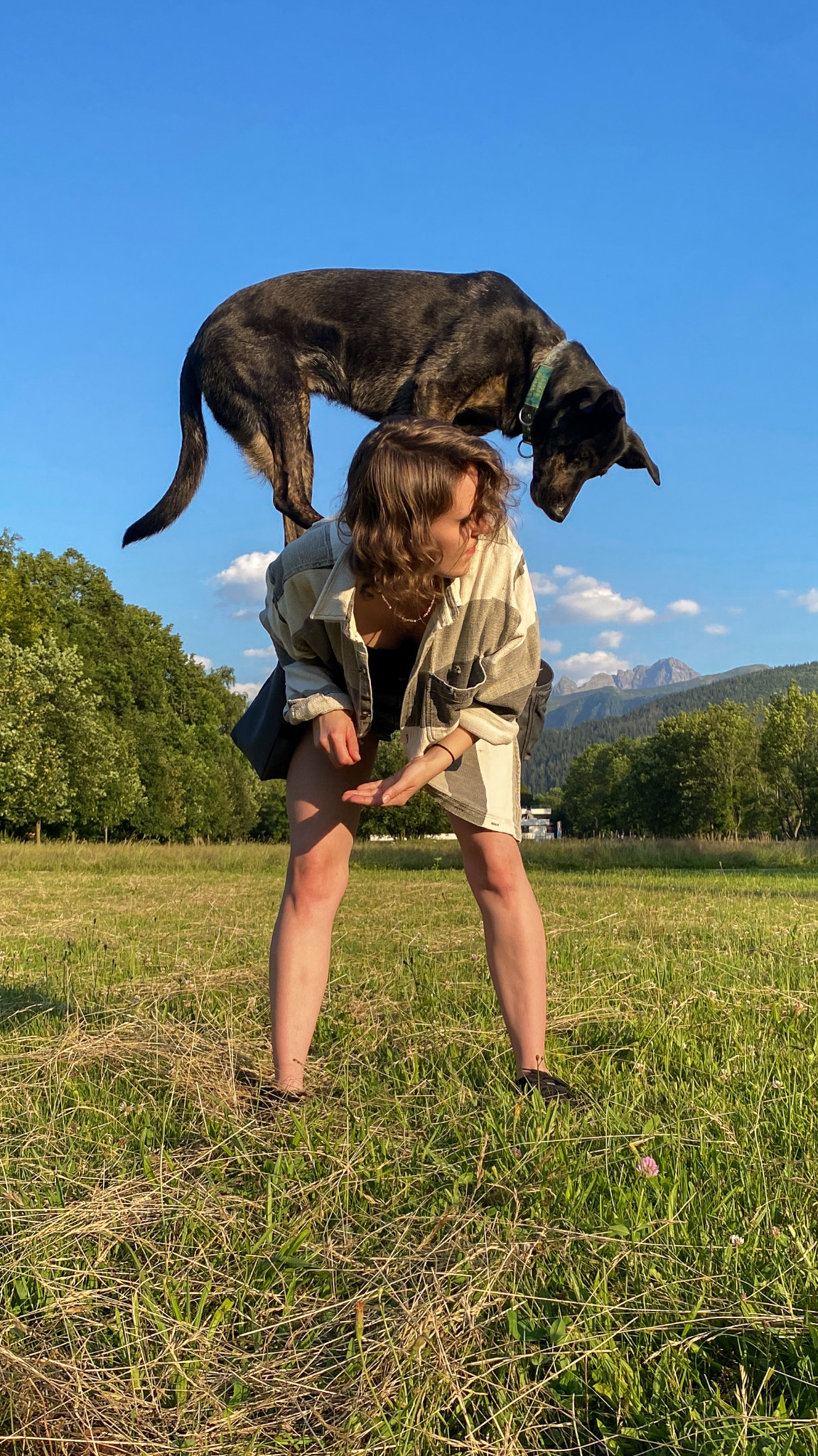Kobieta w kraciastej koszuli z psem stojącym na jej plecach na łące z widokiem na góry w tle.