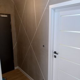 Dawid Mazelewski - Ściana w odcieniach beżu z geometrycznym wzorem białych linii, białe drzwi wewnętrzne z poziomymi przeszkleniami, drewniana podłoga, widoczny fragment odkurzacza.