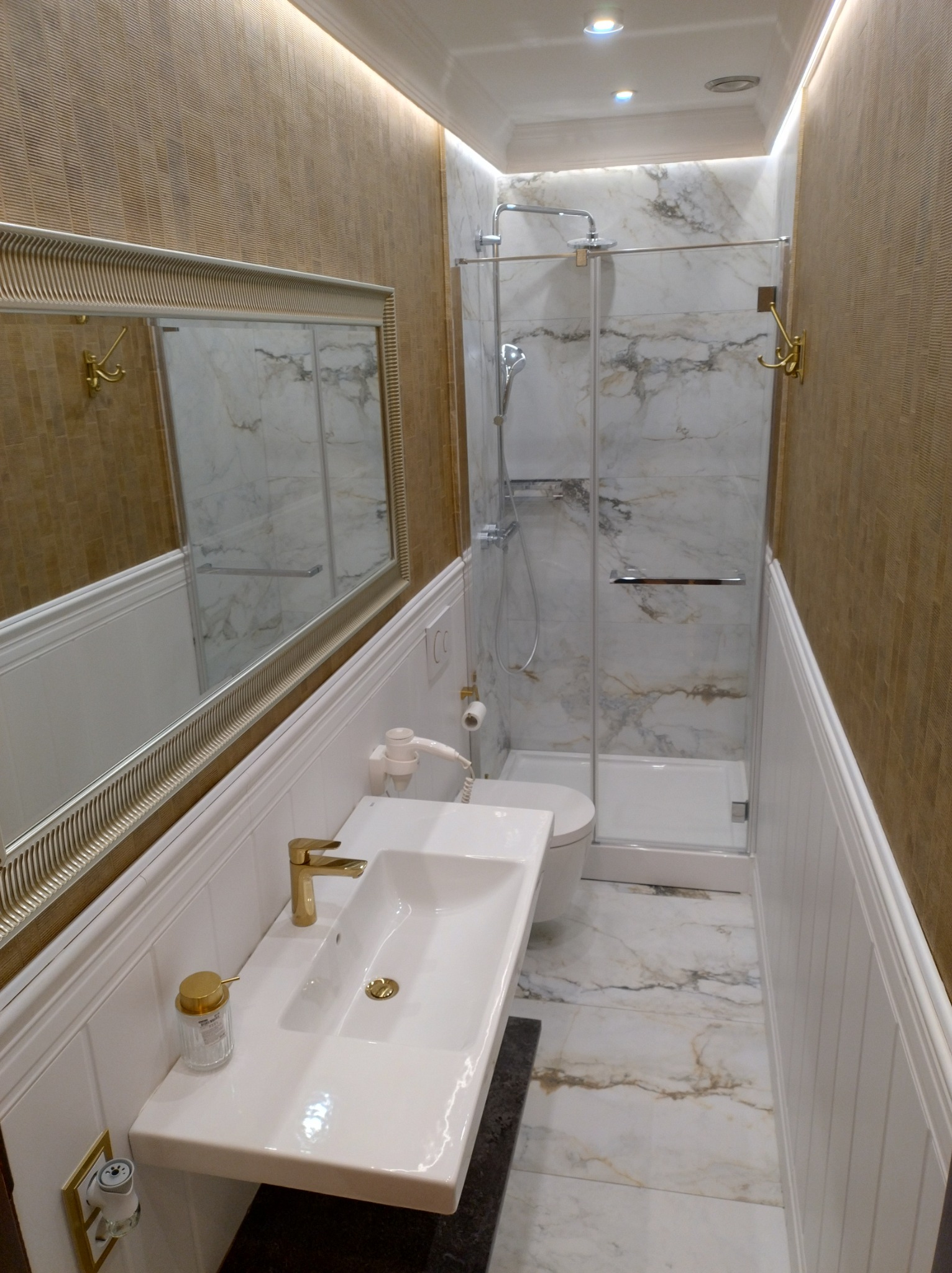 Wąska łazienka z kabiną prysznicową wykończona płytkami imitującymi marmur, złota armatura, lustro w ozdobnej ramie i oświetlenie LED.