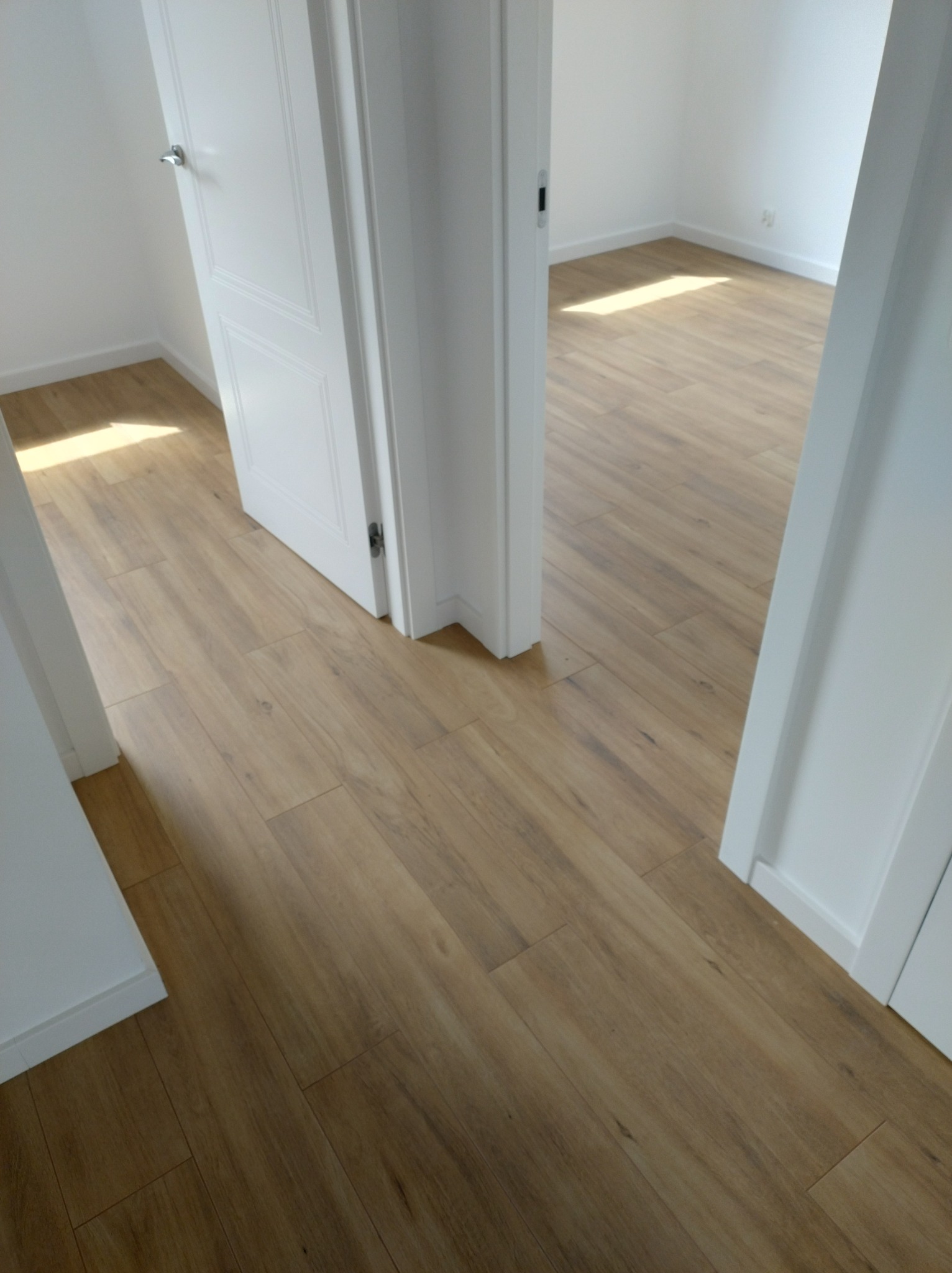 Jasna, drewnopodobna podłoga w nowym pomieszczeniu, widok przez otwarte białe drzwi z chromowaną klamką.