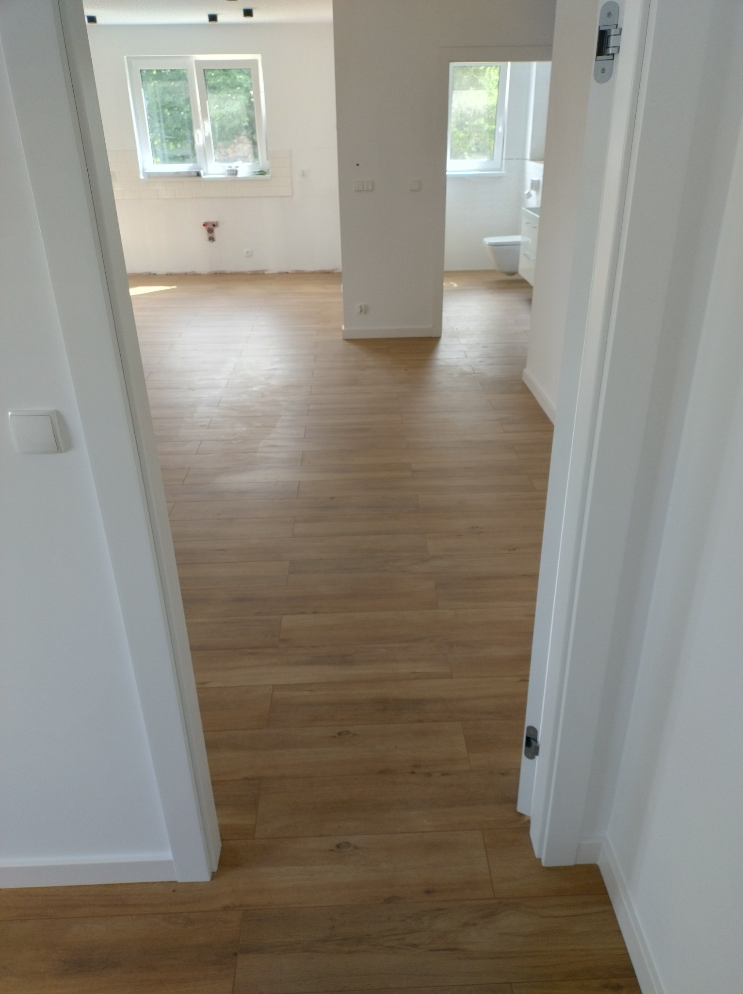 Widok zza uchylonych białych drzwi na jasne, odnowione wnętrze z drewnopodobną podłogą, widoczne okna i fragment łazienki w głębi.