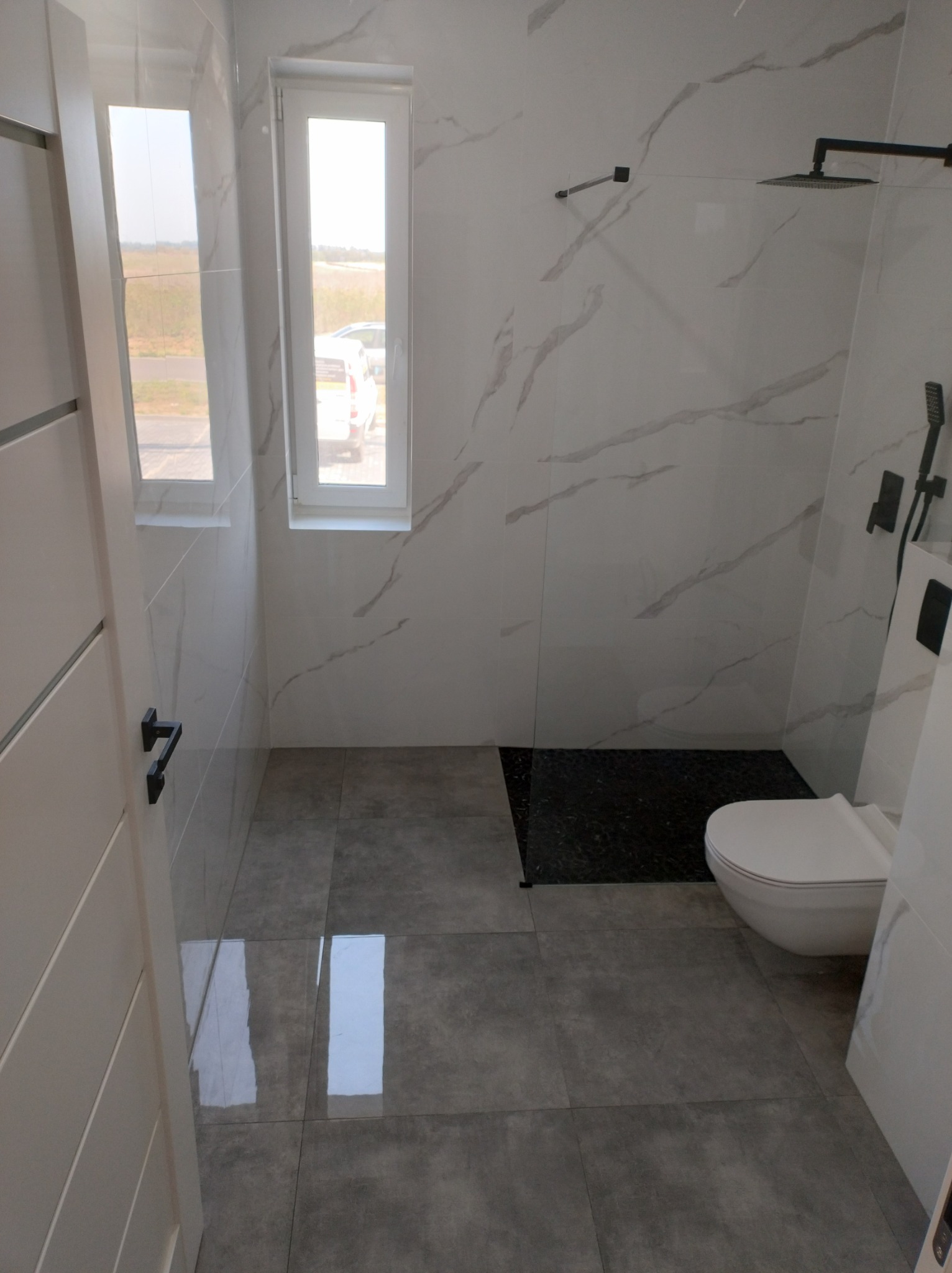 Nowoczesna łazienka z białymi marmurkowymi płytkami ściennymi, szarą podłogą z połyskiem, czarnym brodzikiem i białym WC, widok zza uchylonych drzwi.