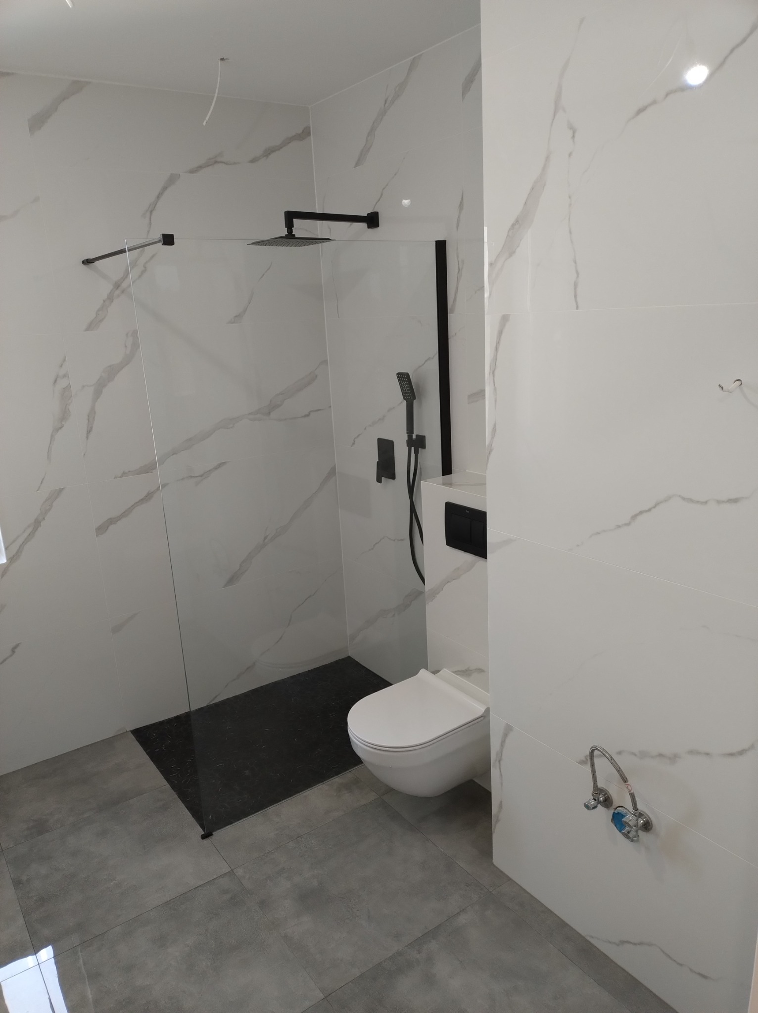 Nowoczesna łazienka z kabiną prysznicową typu walk-in z czarną armaturą i deszczownicą, białą toaletą podwieszaną oraz białymi płytkami imitującymi marmur na ścianach i szarymi płytkami na podłodze.