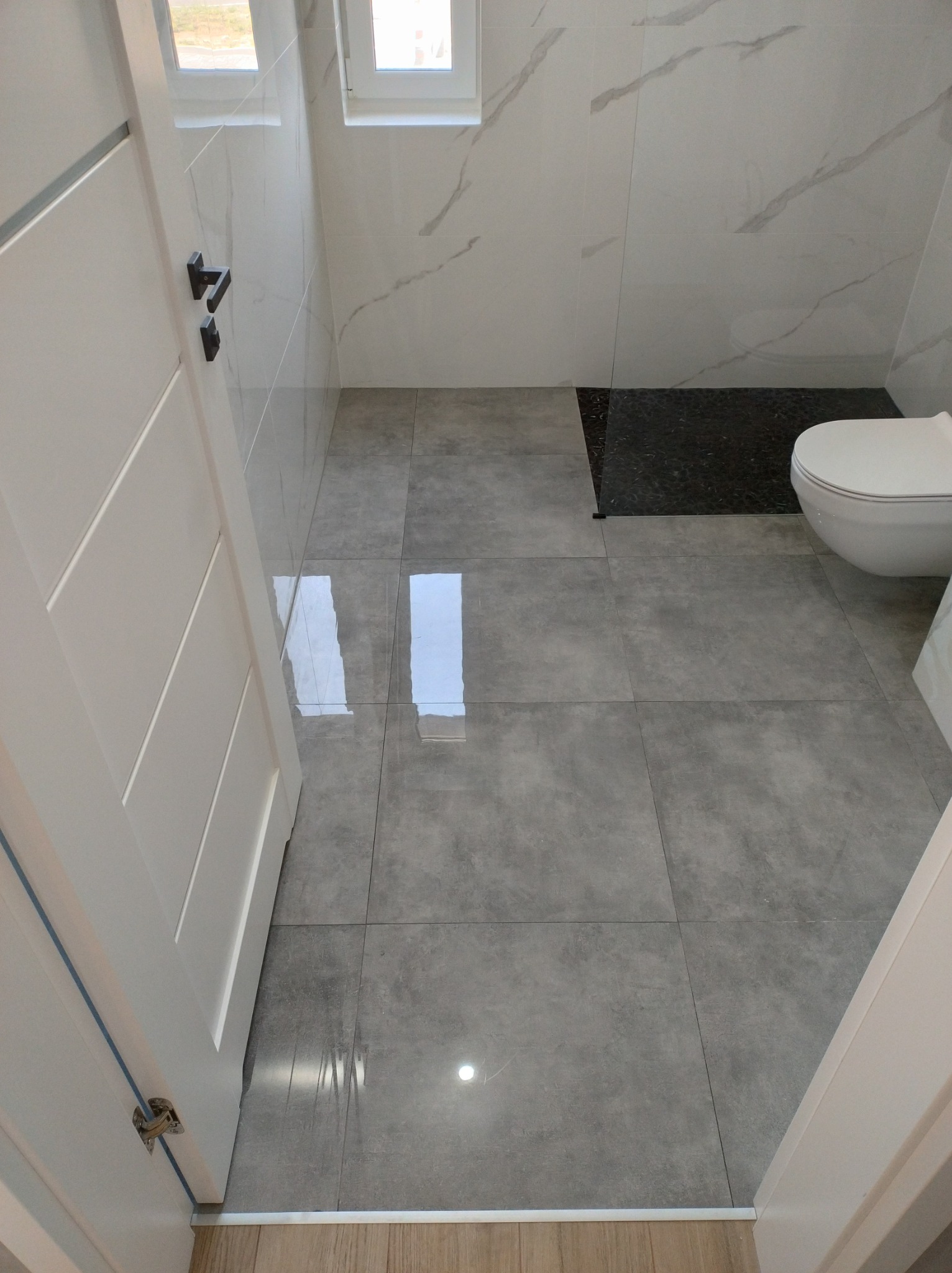 Nowoczesna łazienka z szarymi płytkami podłogowymi, białymi marmurkowymi płytkami ściennymi i czarną strefą prysznicową, widok zza otwartych białych drzwi.