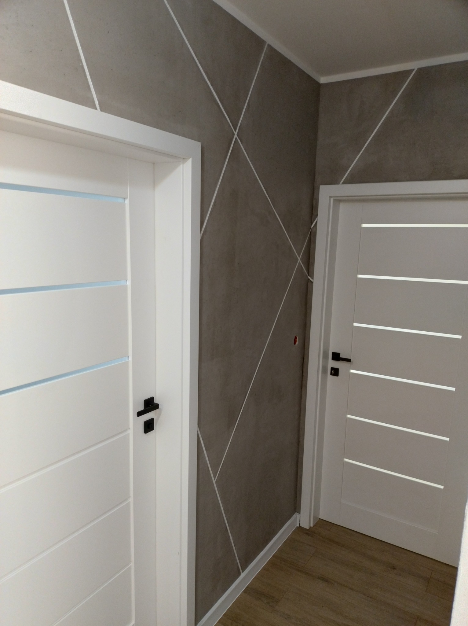 Szara ściana z dekoracyjnymi białymi liniami w jodełkę, dwie białe drzwi z poziomymi wgłębieniami i czarnymi klamkami, drewniana podłoga