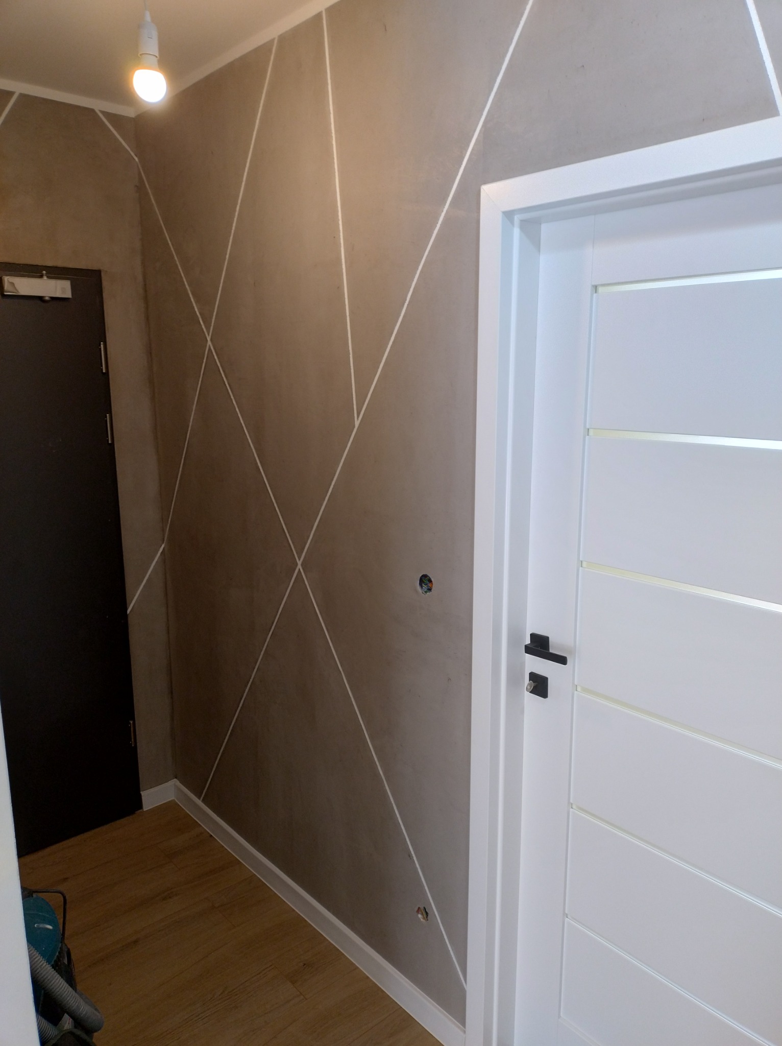 Ściana w odcieniach beżu z geometrycznym wzorem białych linii, białe drzwi wewnętrzne z poziomymi przeszkleniami, drewniana podłoga, widoczny fragment odkurzacza.