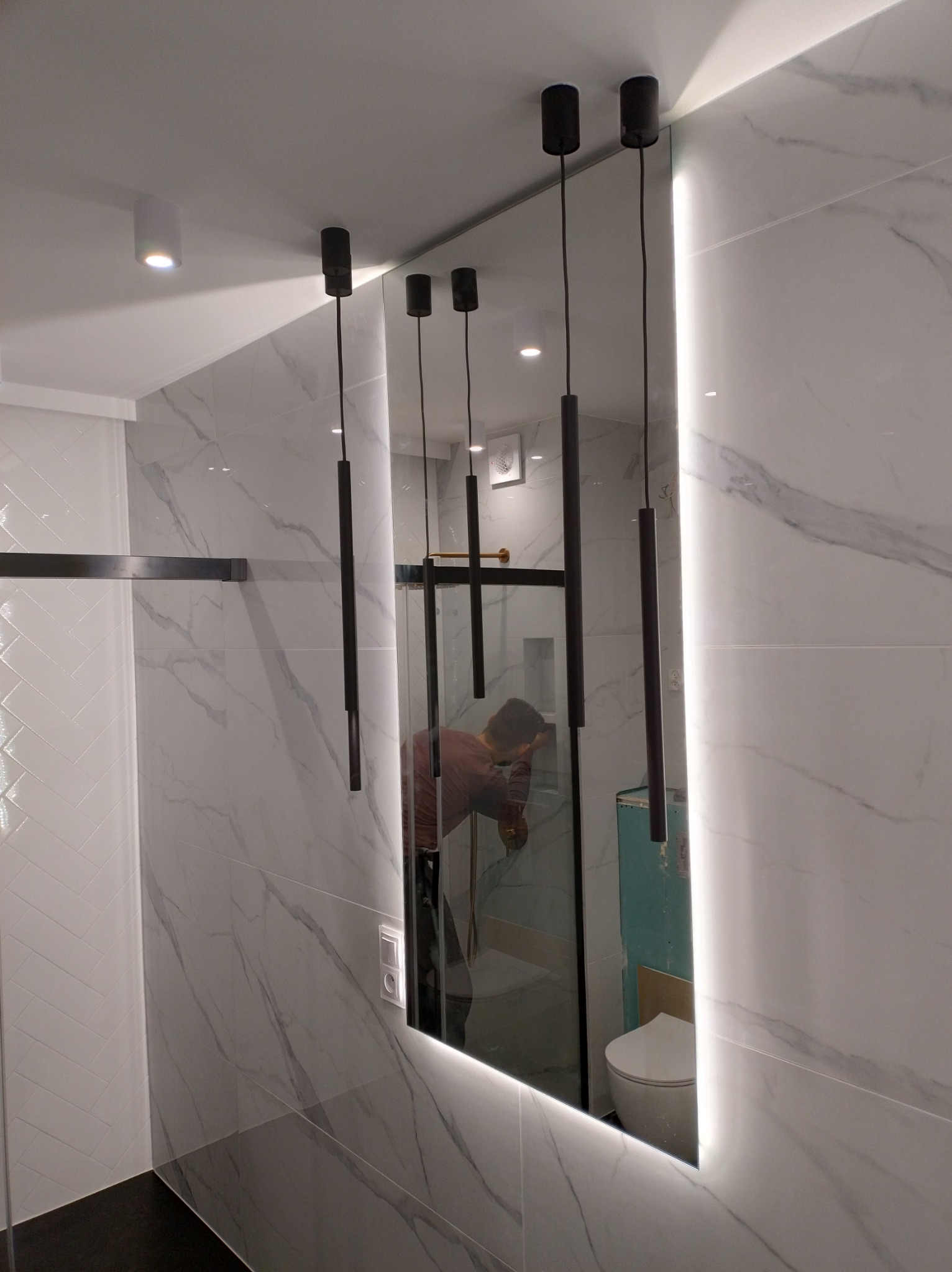 Nowoczesna łazienka z marmurowymi płytkami, podświetlanym lustrem i wiszącymi lampami, w odbiciu widoczna osoba pracująca nad instalacją kabiny prysznicowej.