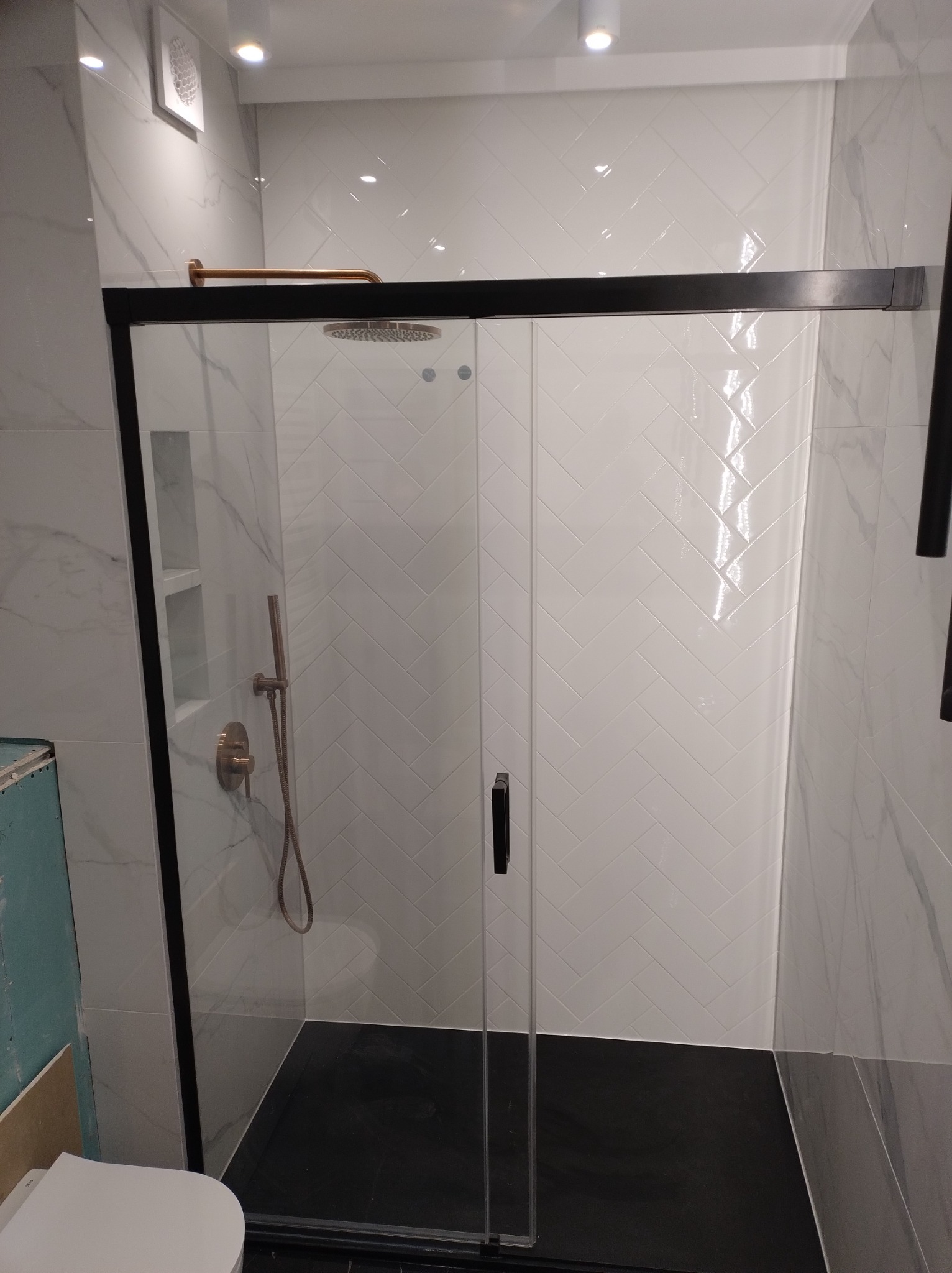 Nowoczesna kabina prysznicowa z czarną ramą, deszczownicą i baterią w kolorze miedzi, białe płytki ułożone w jodełkę na ścianie, czarna posadzka.