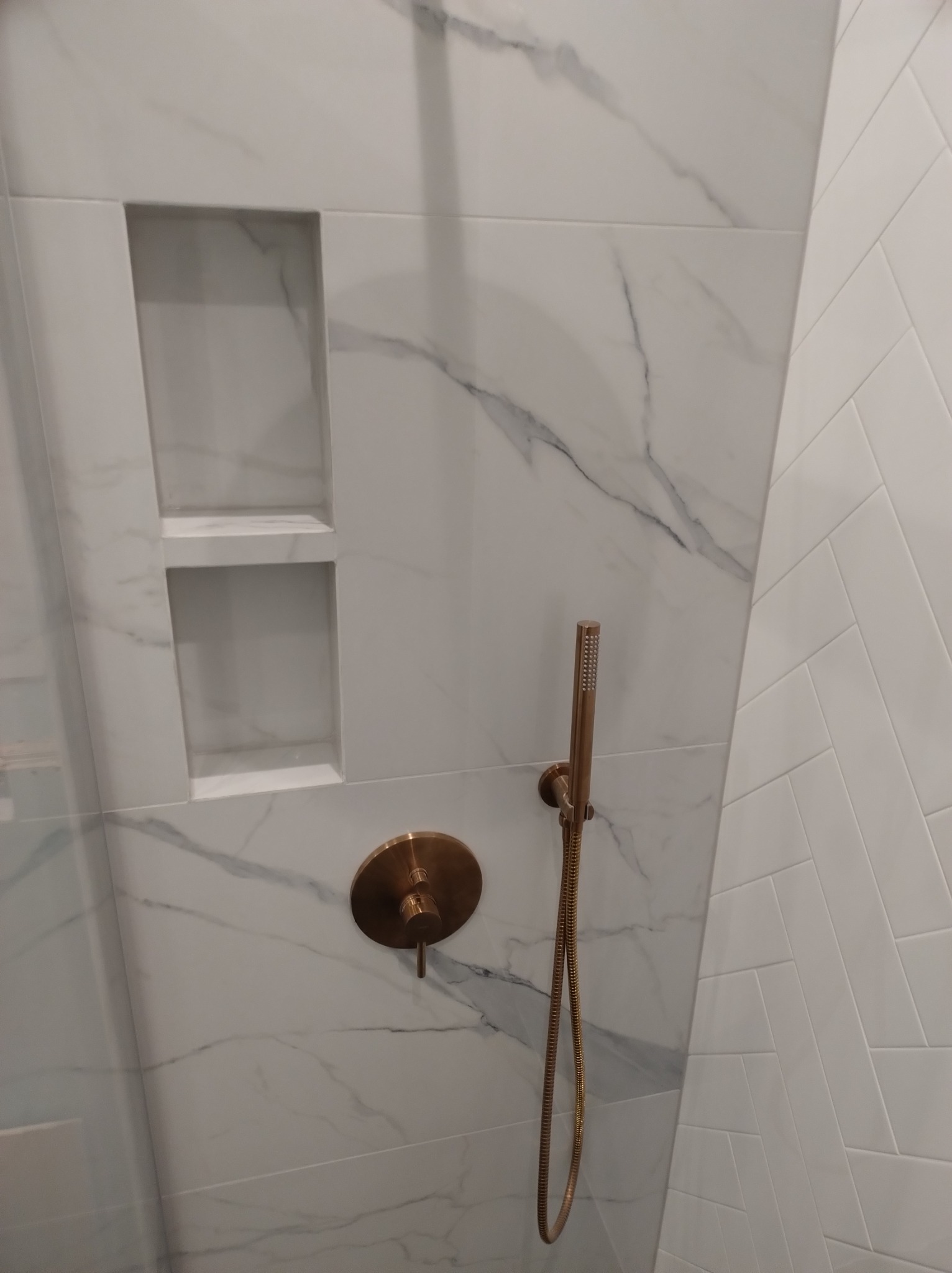 Wykończona łazienka: Białe płytki imitujące marmur z szarymi żyłami, wnęka z półkami, mosiężna bateria i deszczownica, ściana z białych płytek ułożonych w jodełkę.