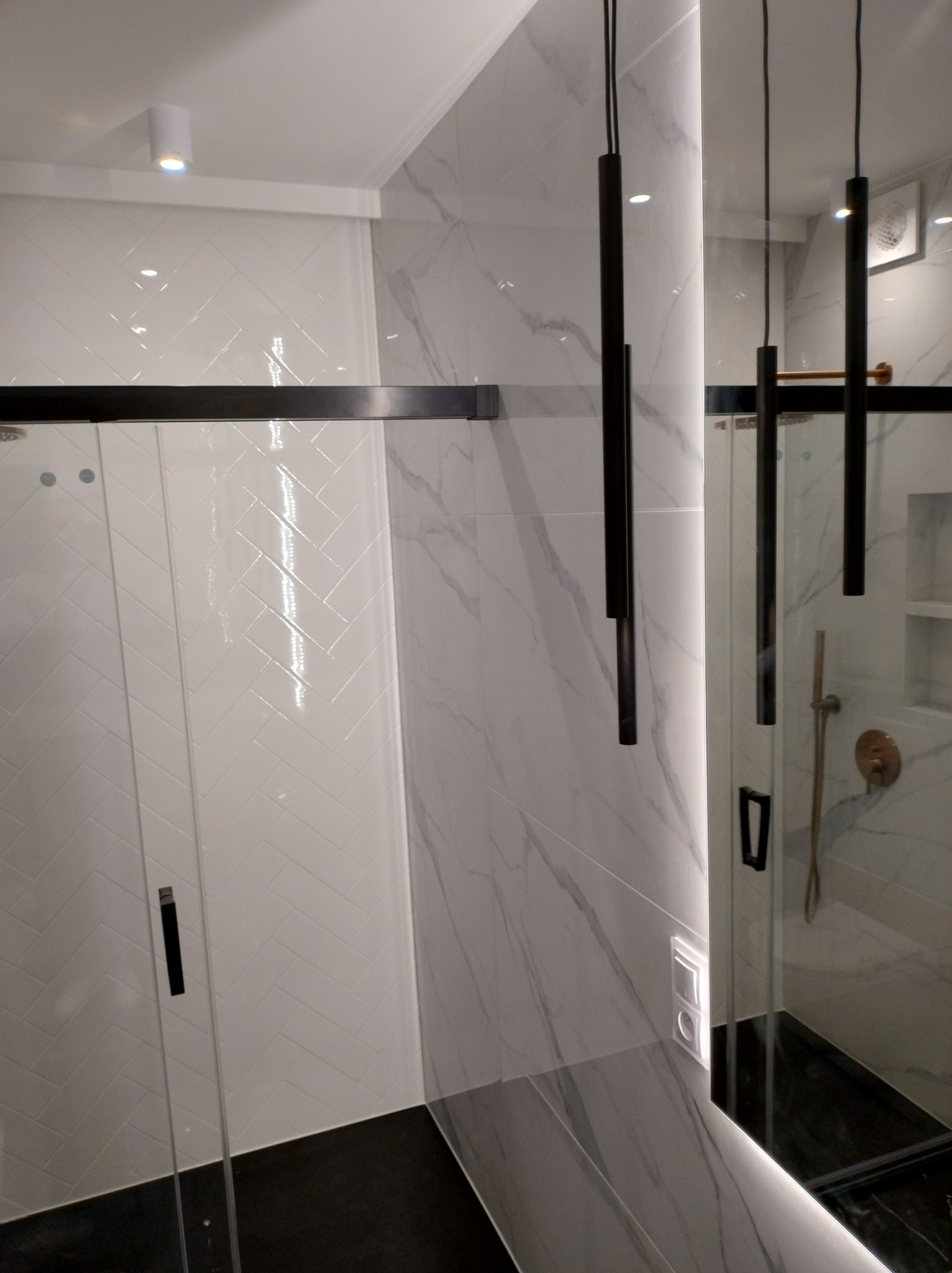 Nowoczesna łazienka z kabiną prysznicową, białymi płytkami ułożonymi w jodełkę, marmurowymi panelami ściennymi i minimalistycznymi lampami wiszącymi.
