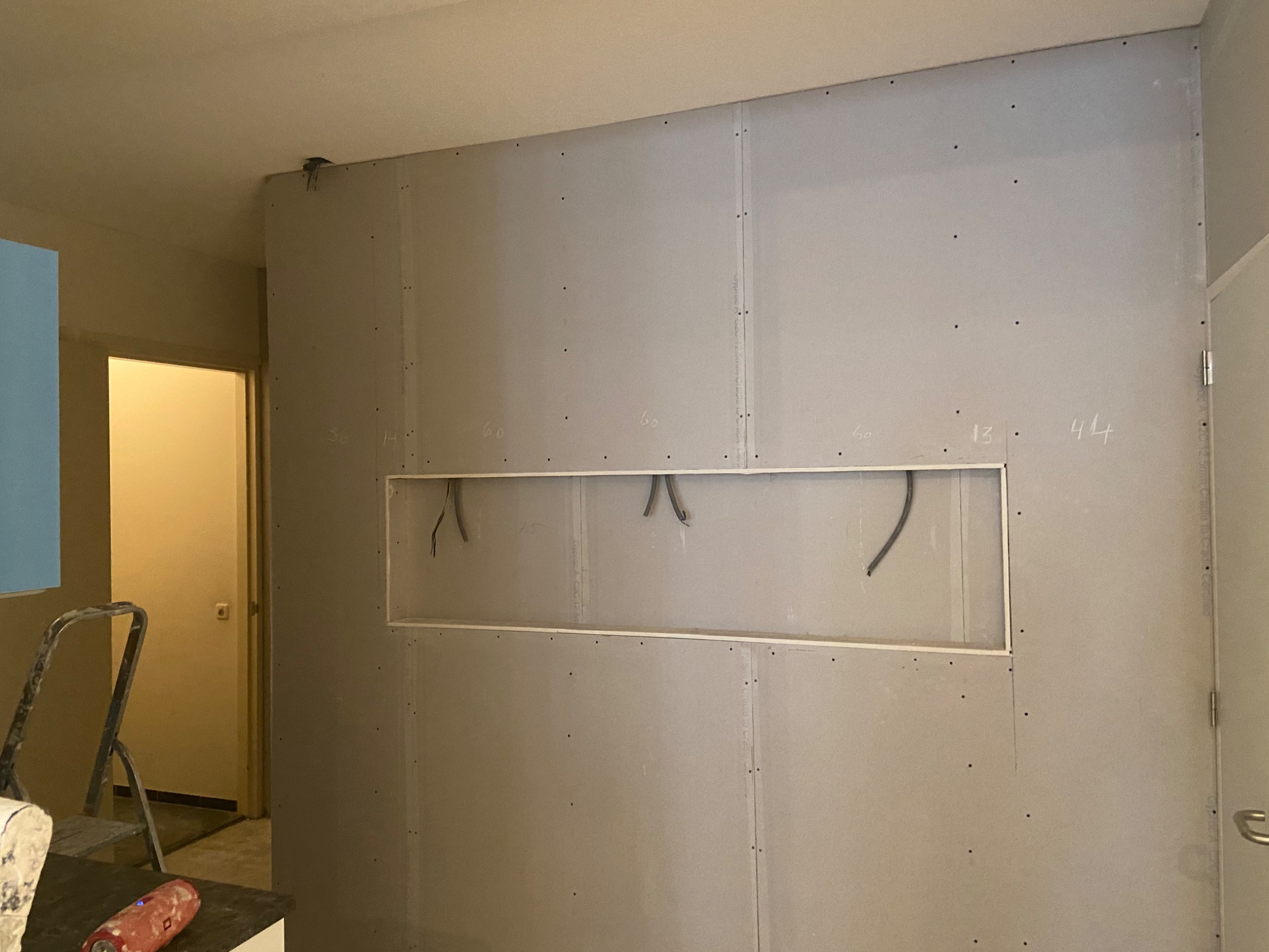 Ściana z płyt gipsowo-kartonowych w trakcie montażu, widoczne otwory na instalacje, surowe wykończenie, ślady pomiarów ołówkiem, drabina malarska w tle.