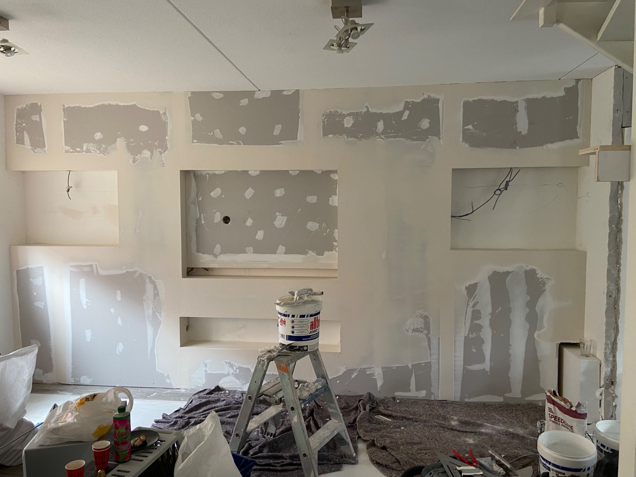 Ściana w trakcie wyrównywania gładzią szpachlową, widoczne ślady po łataniu ubytków, drabina z wiadrem gładzi na tle przygotowanego pomieszczenia.