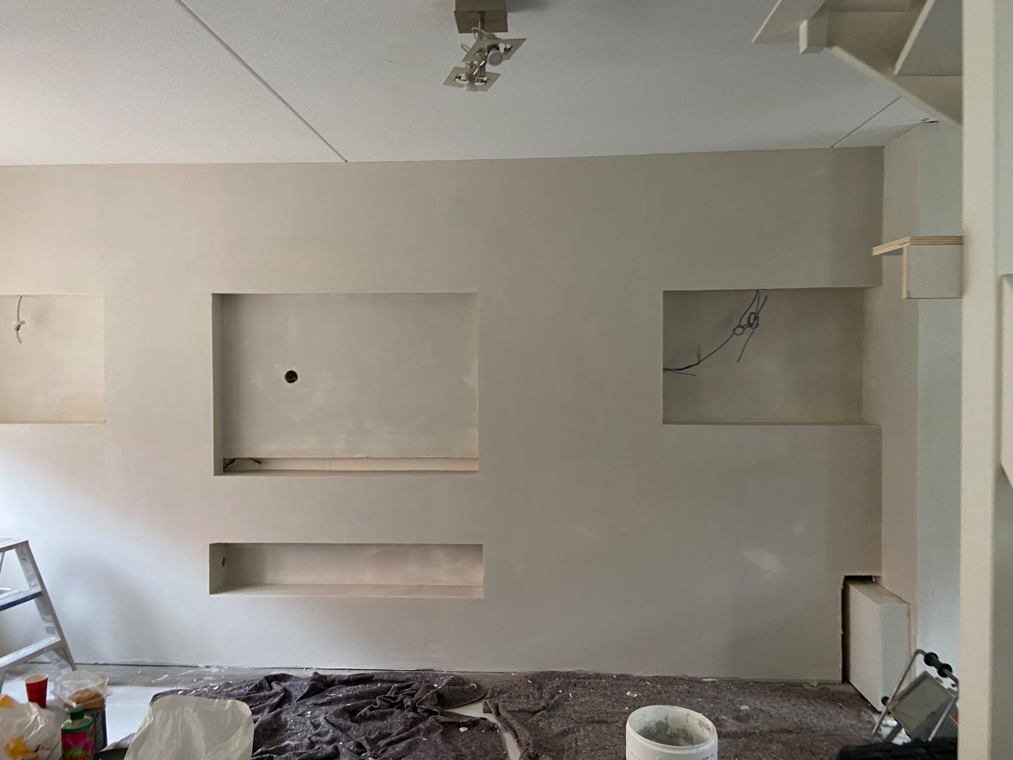 Ściana w trakcie prac wykończeniowych z wnękami, widoczne ślady po gładzi, drabina i akcesoria malarskie na podłodze zabezpieczonej folią.