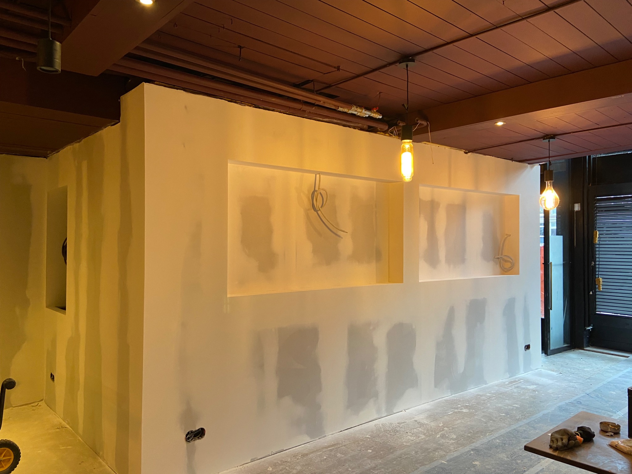 Ściana w trakcie wygładzania z wnękami oświetlonymi żarówkami Edison, widoczne ślady szpachli i instalacje elektryczne, sufit z drewnianych belek pomalowanych na brązowo.