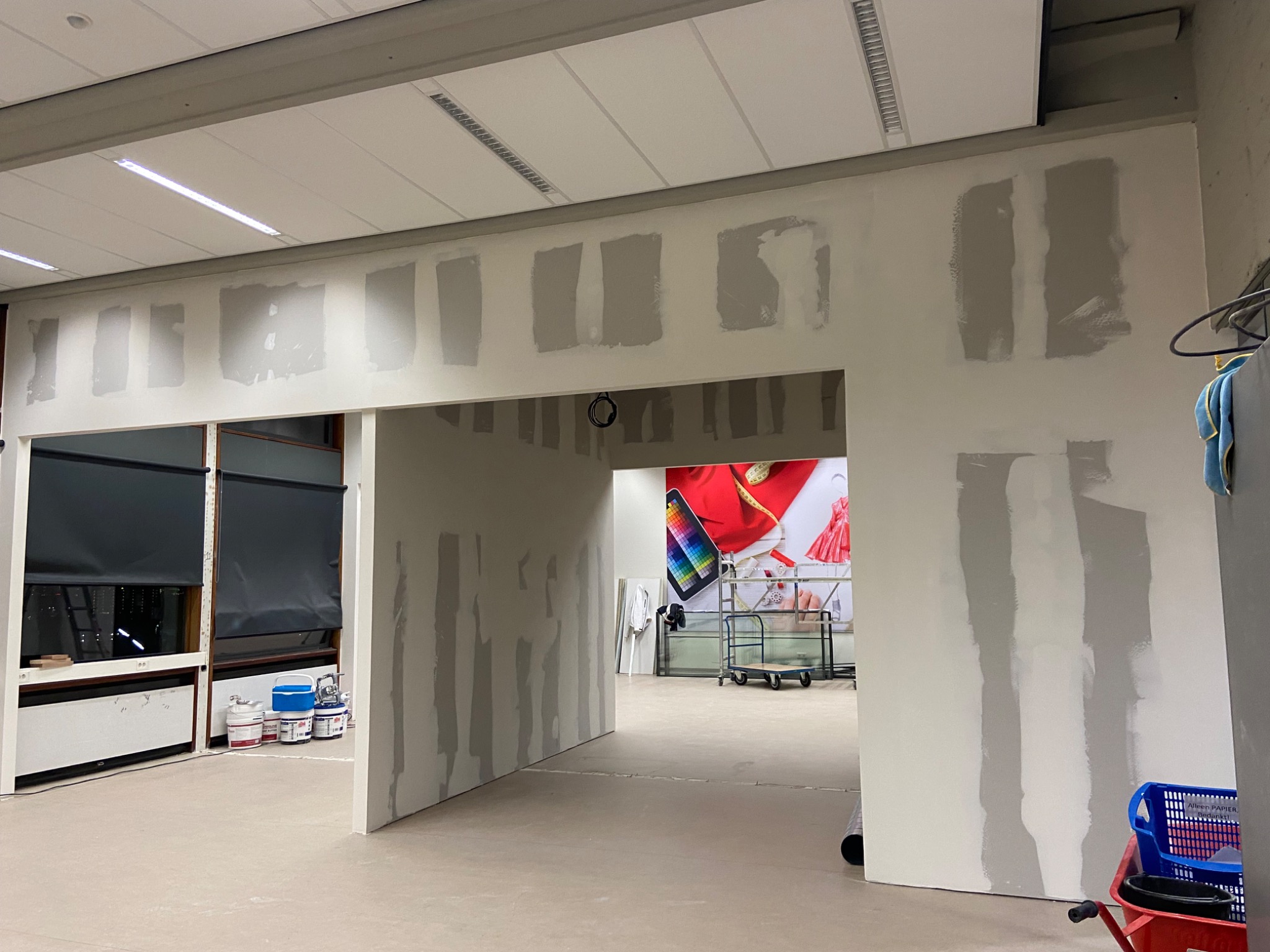 Fragment ściany w trakcie prac wykończeniowych z widocznymi pionowymi pasami nałożonej gładzi, w tle widoczne puszki z farbą i elementy wyposażenia wnętrza sklepu odzieżowego.