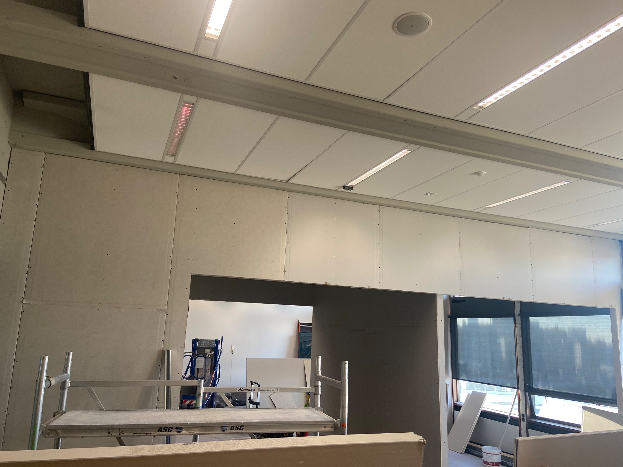 Wnętrze w trakcie prac wykończeniowych, widoczne płyty gipsowo-kartonowe na ścianach, sufit podwieszany z oświetleniem LED, rusztowanie aluminiowe oraz fragment okna z roletami.