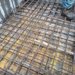 Zbrojenie fundamentu z siatki stalowej na żółtym szalunku, widoczne buty robocze i fragment betonowej ściany.