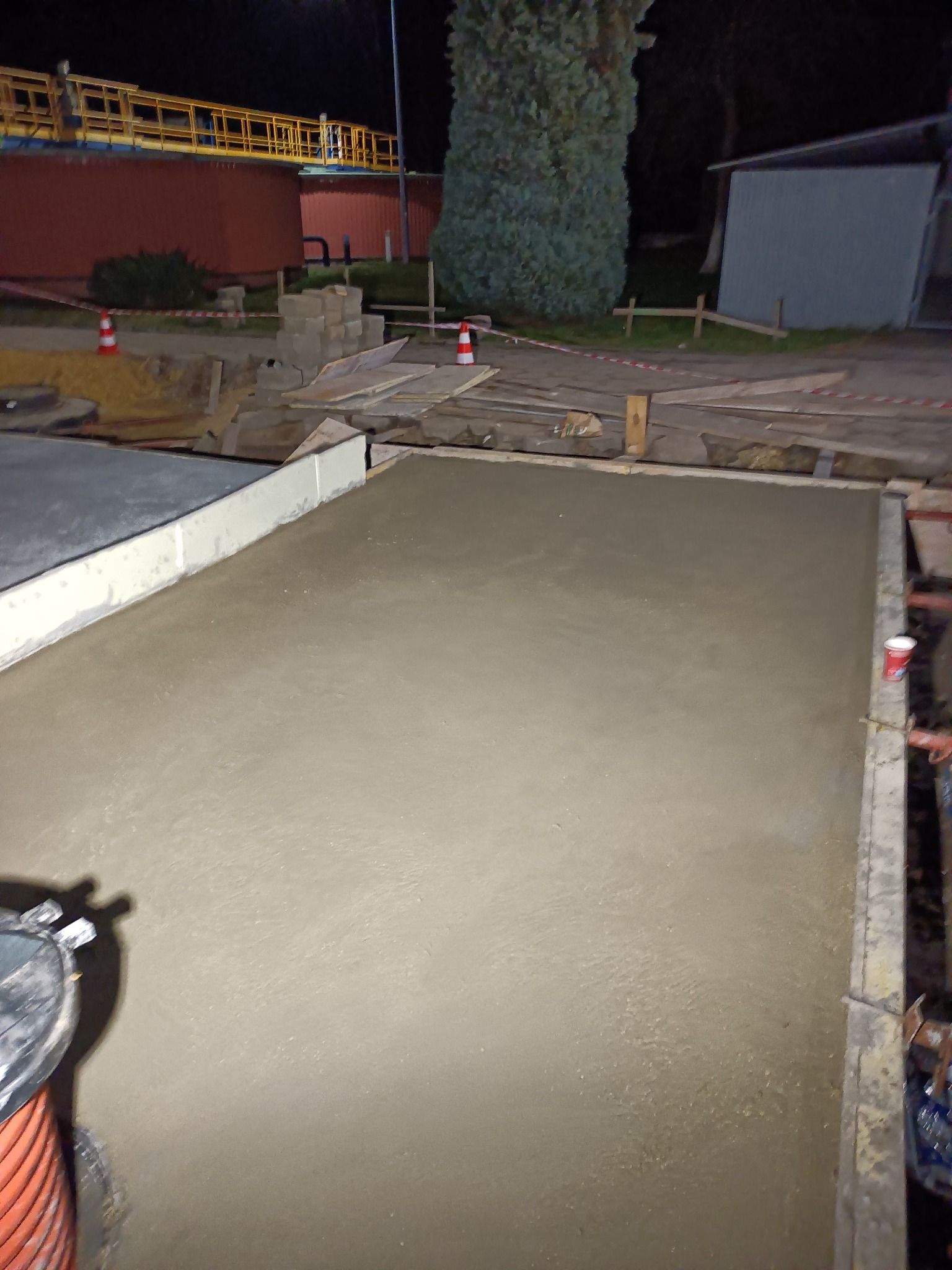 Świeżo wylana, gładka betonowa płyta fundamentowa w szalunku, widoczne elementy konstrukcyjne i kubek po napoju, na tle nocnego krajobrazu przemysłowego.