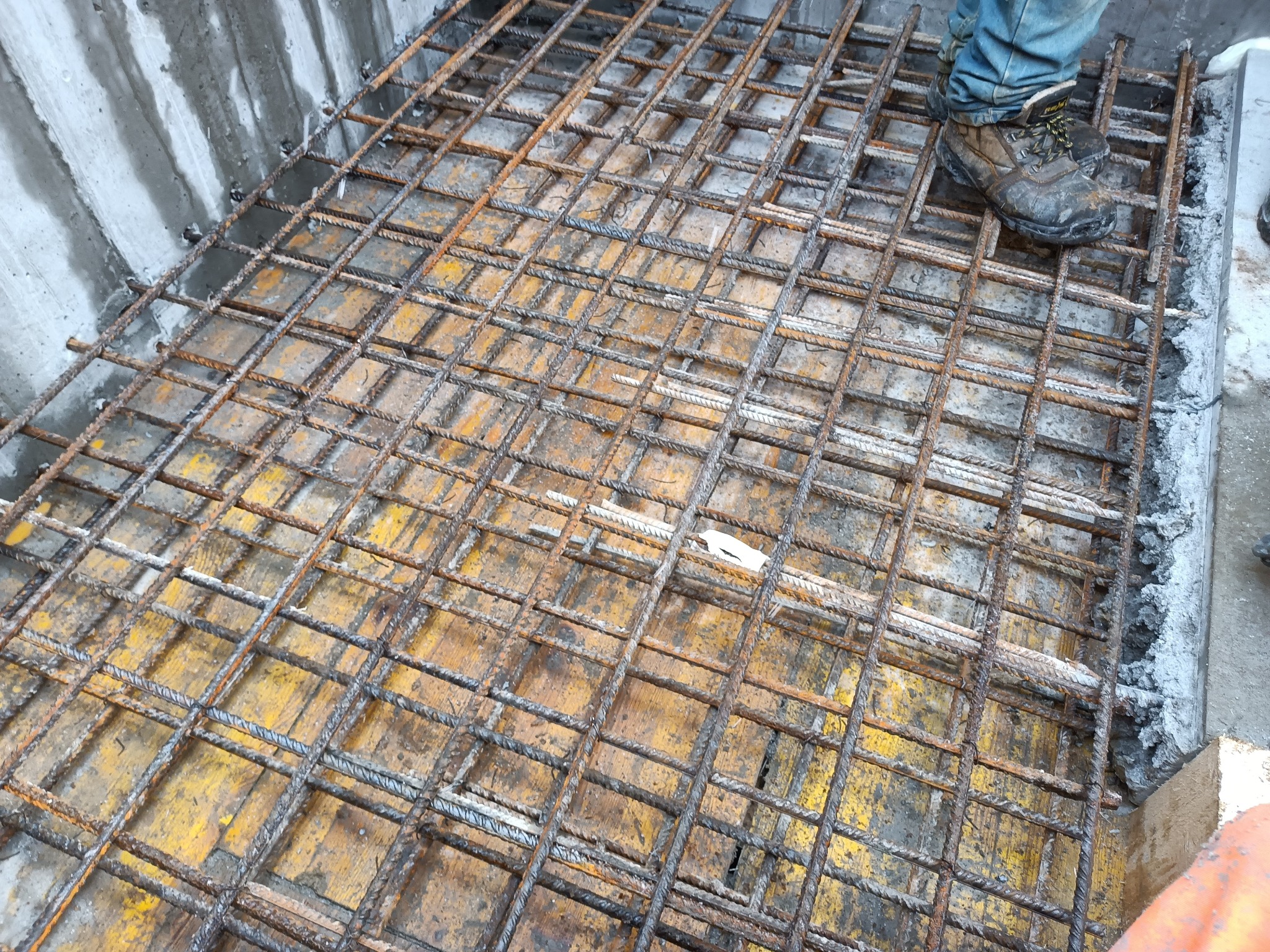 Zbrojenie fundamentu z siatki stalowej na żółtym szalunku, widoczne buty robocze i fragment betonowej ściany.